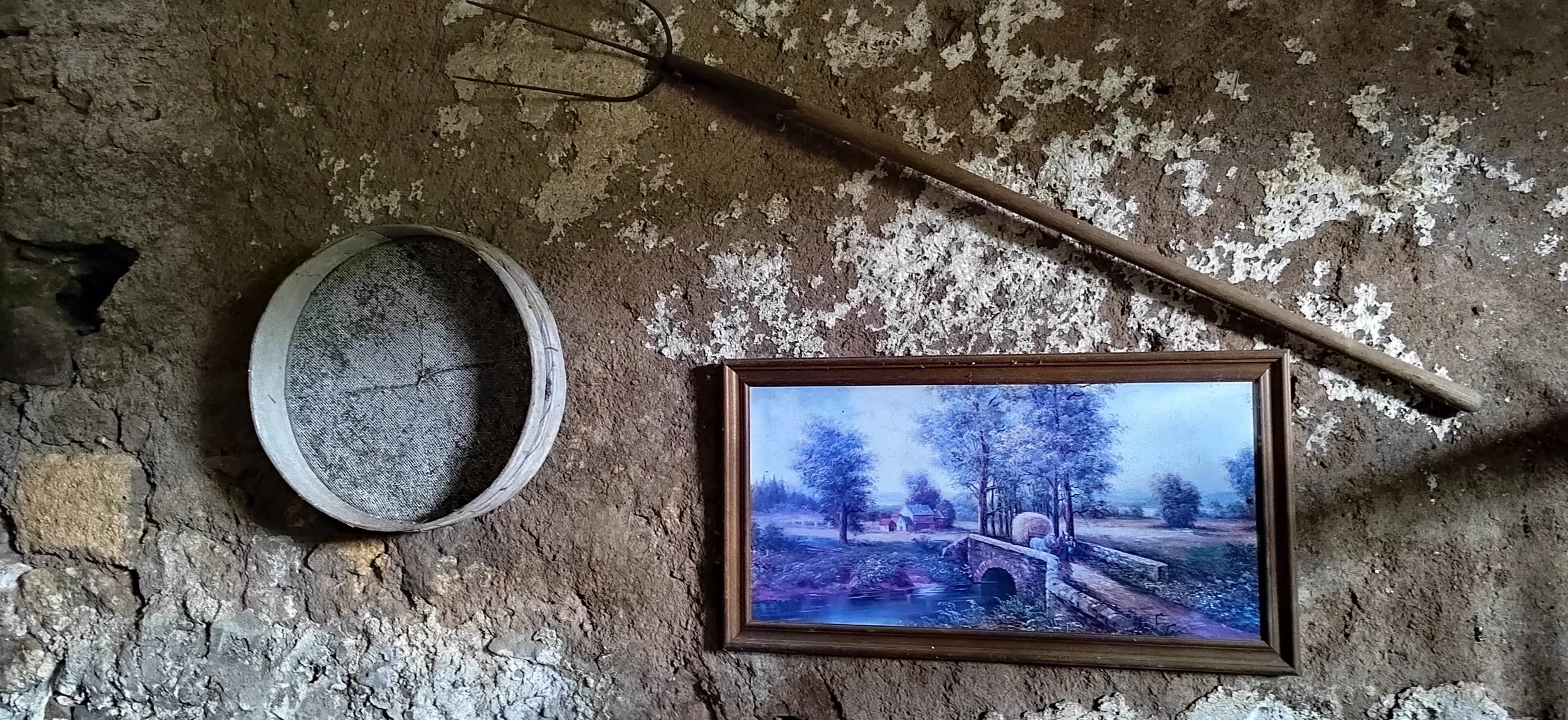 Ein altes Gemälde zeigt eine ländliche Szenerie mit Bäumen, einem Fluss, einem kleinen Boot, einem Haus und einer Brücke, alles in einem Holzrahmen. Neben dem Gemälde liegt eine runde Kiefernzapfen und ein langer Holzstab auf getrocknetem Boden.