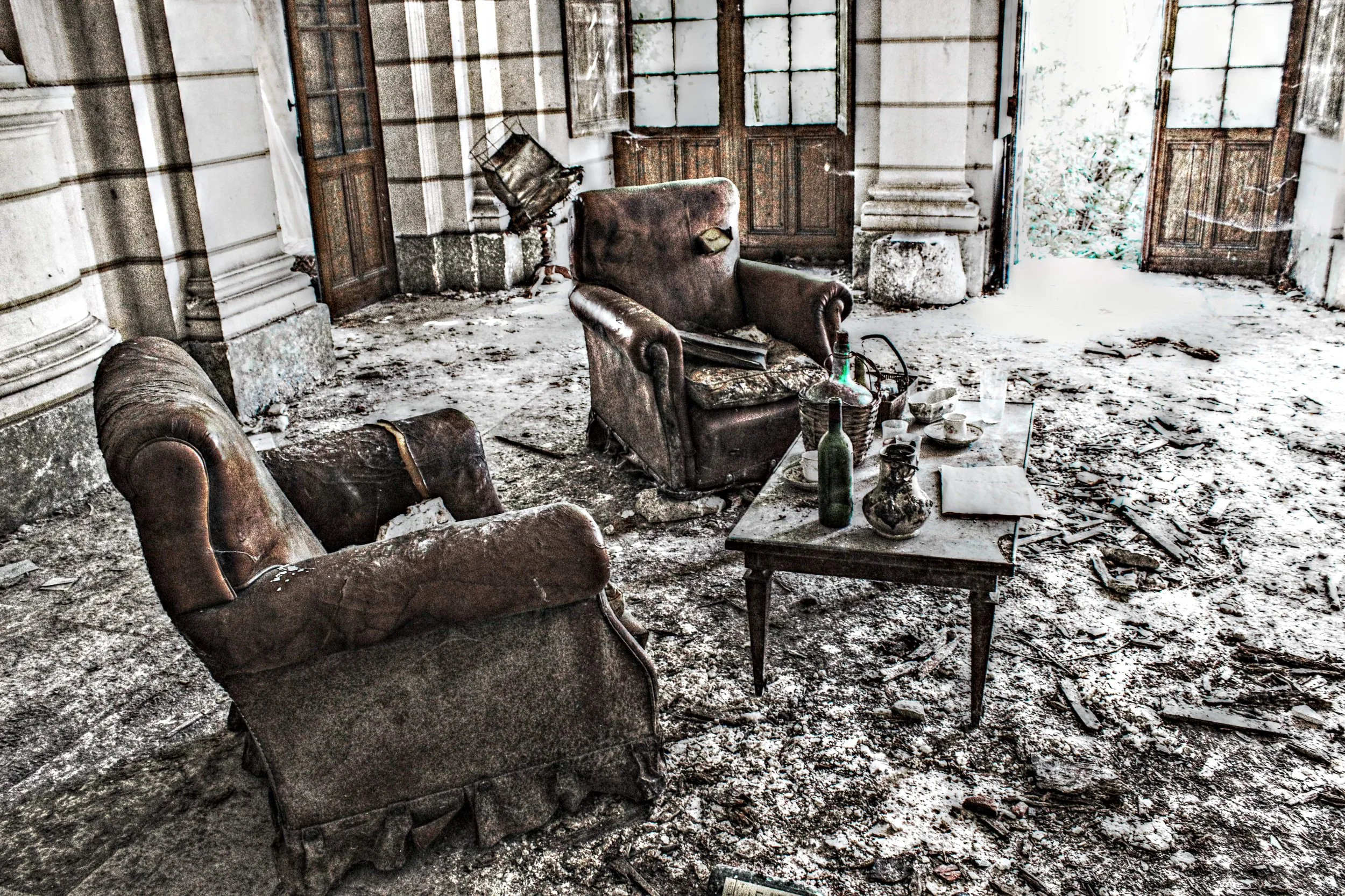 Verwitterte, abgenutzte, alte Ledersessel und ein Couchtisch in einem vernachlässigten Raum mit staubigem Boden und zerbrochenen Fenstern.