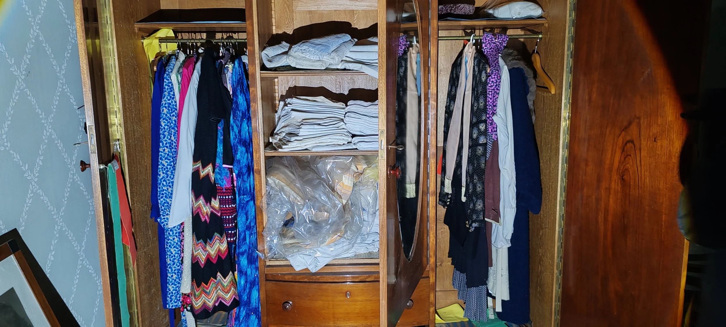 Offene Holzschrank mit Kleidung, darunter eine kleine Schublade, im Hintergrund ein blauer Wandteppich mit weißen Mustern.