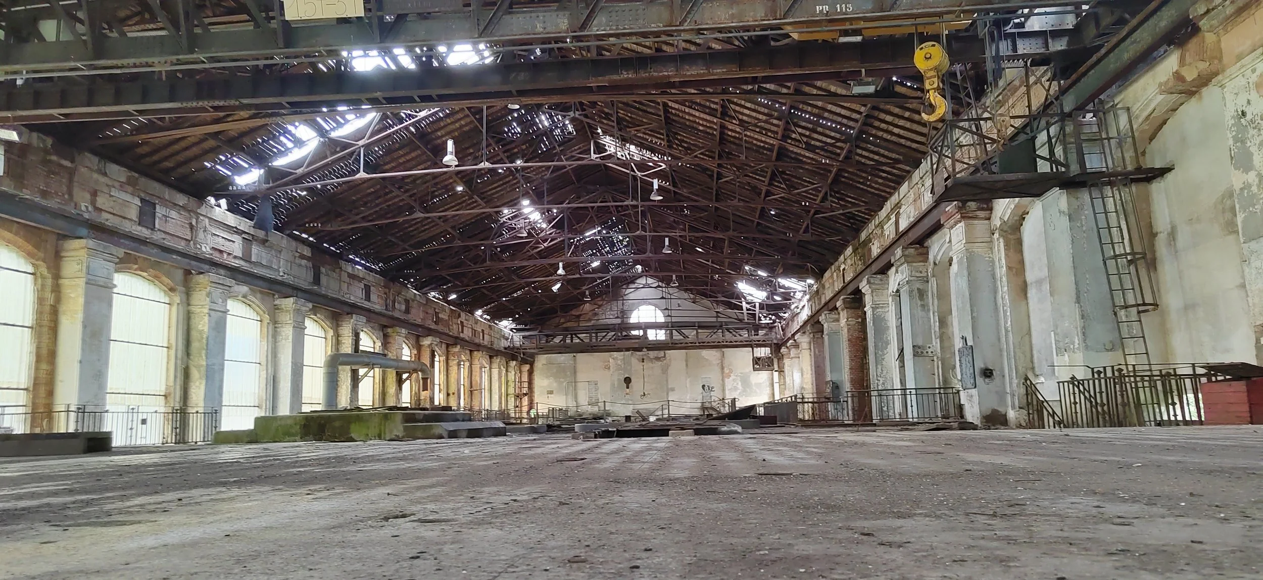 Innenansicht eines alten, verfallenen Industriegebäudes mit hohem Dach und zerbrochenen Fenstern, darunter eine alte, verfallene Etage, auf dem Boden liegen lose Dielen und Schutt.