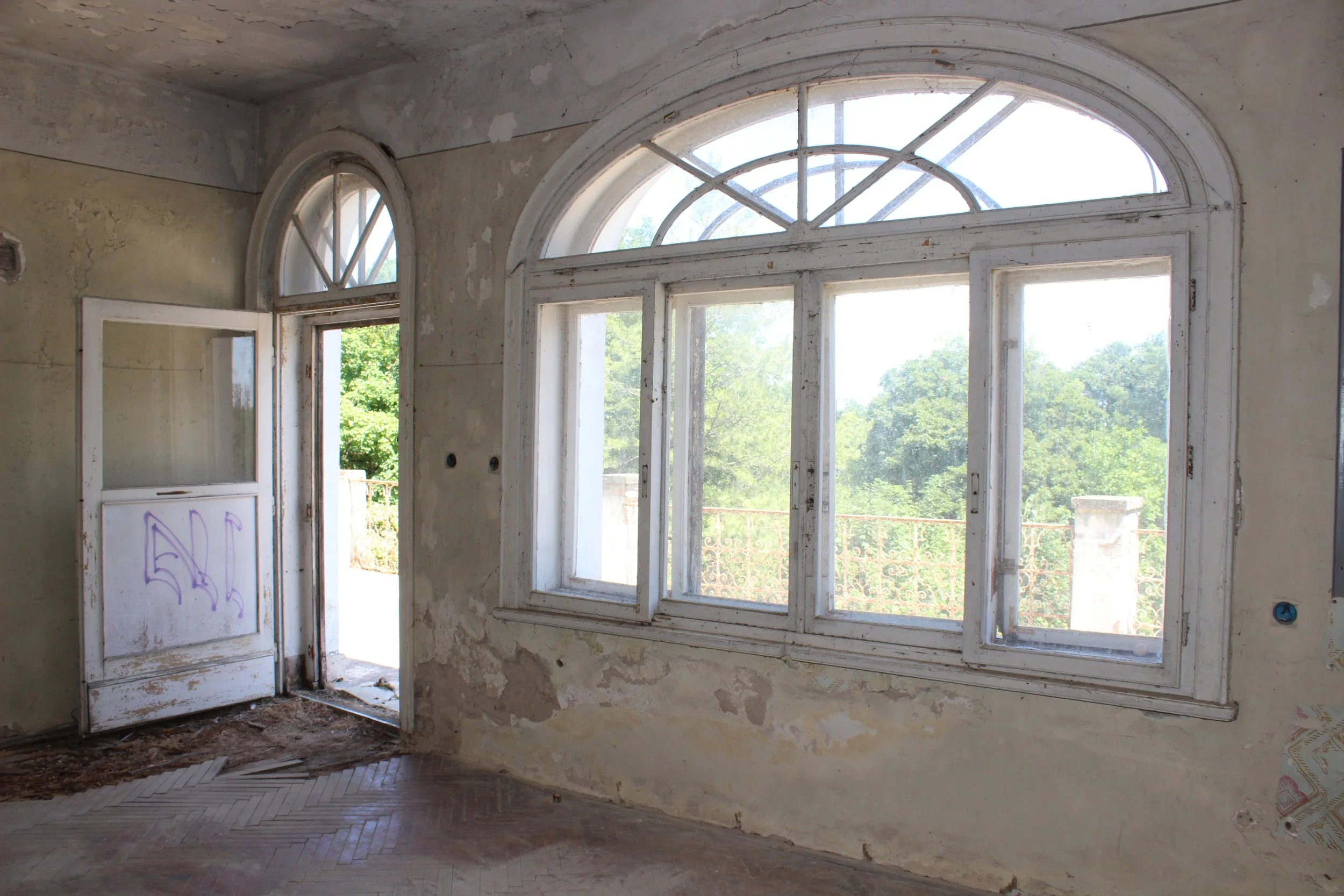 Innenansicht eines veralteten, ungepflegten Zimmers mit großen, alten Fenstern und einem offenen Tür. Das Zimmer ist verlassen und hat abgenutzte Wände mit Flecken und Rissen, sowie einen Boden mit abgenutztem Parkett.