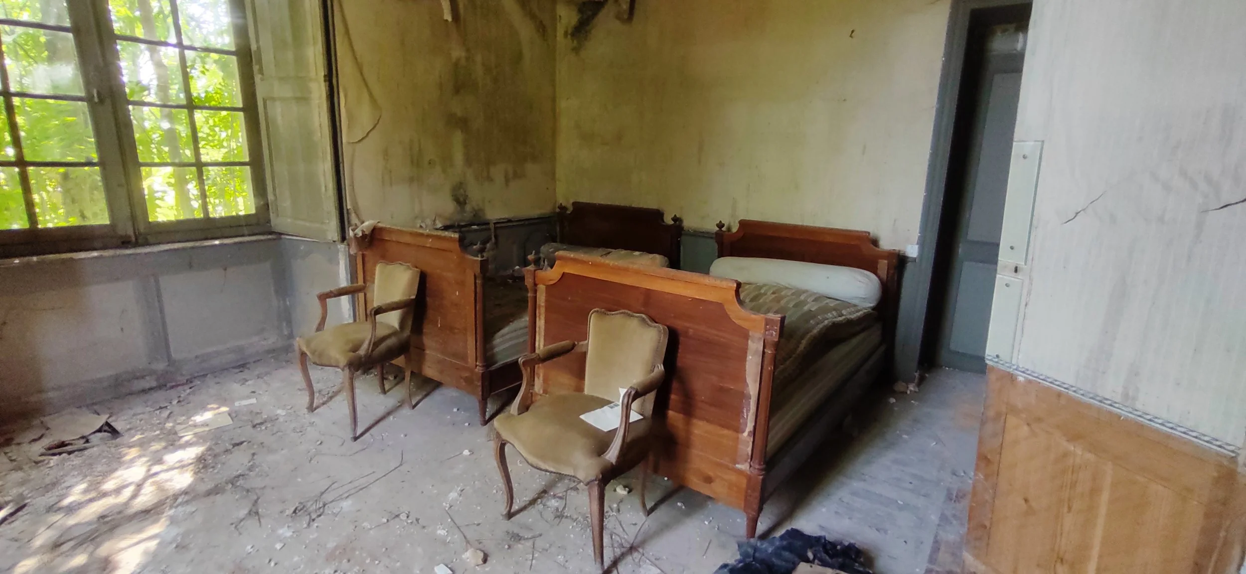 Alte, verlassene Schlafzimmer mit vier Holzbetten, zwei armlehnstühle und schmutzigen, beschädigten Boden, überwucherten Fenster und verwitterten Wänden.