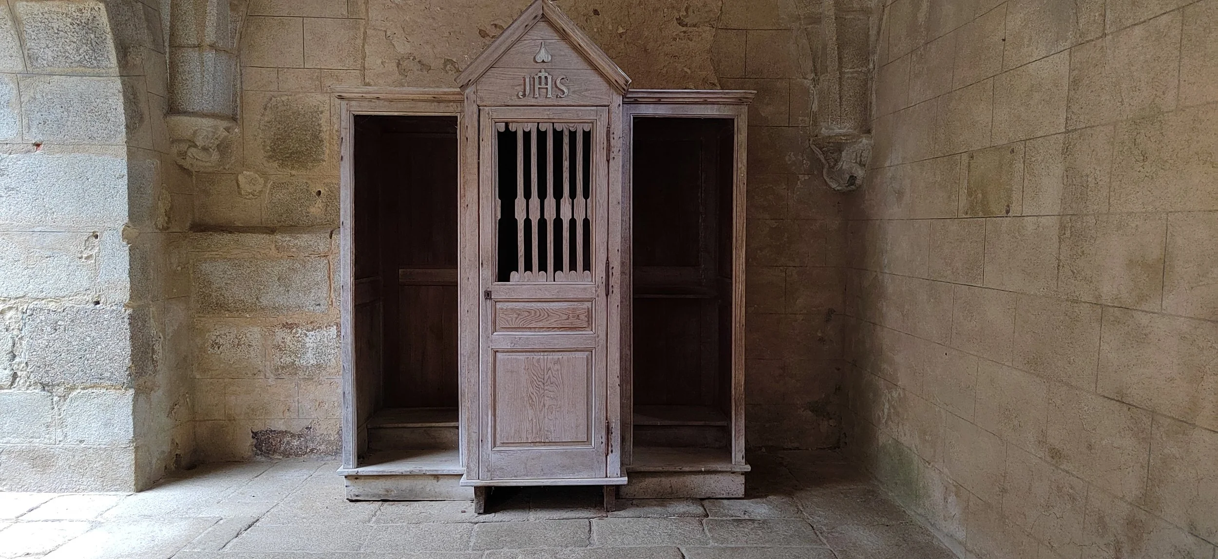 Ein alter, weißlicher Holz-Kasten in einer steinernen Kirche, gekennzeichnet mit den Buchstaben 'JHS' oben, steht an einer rechten Wand auf einem Steinboden.