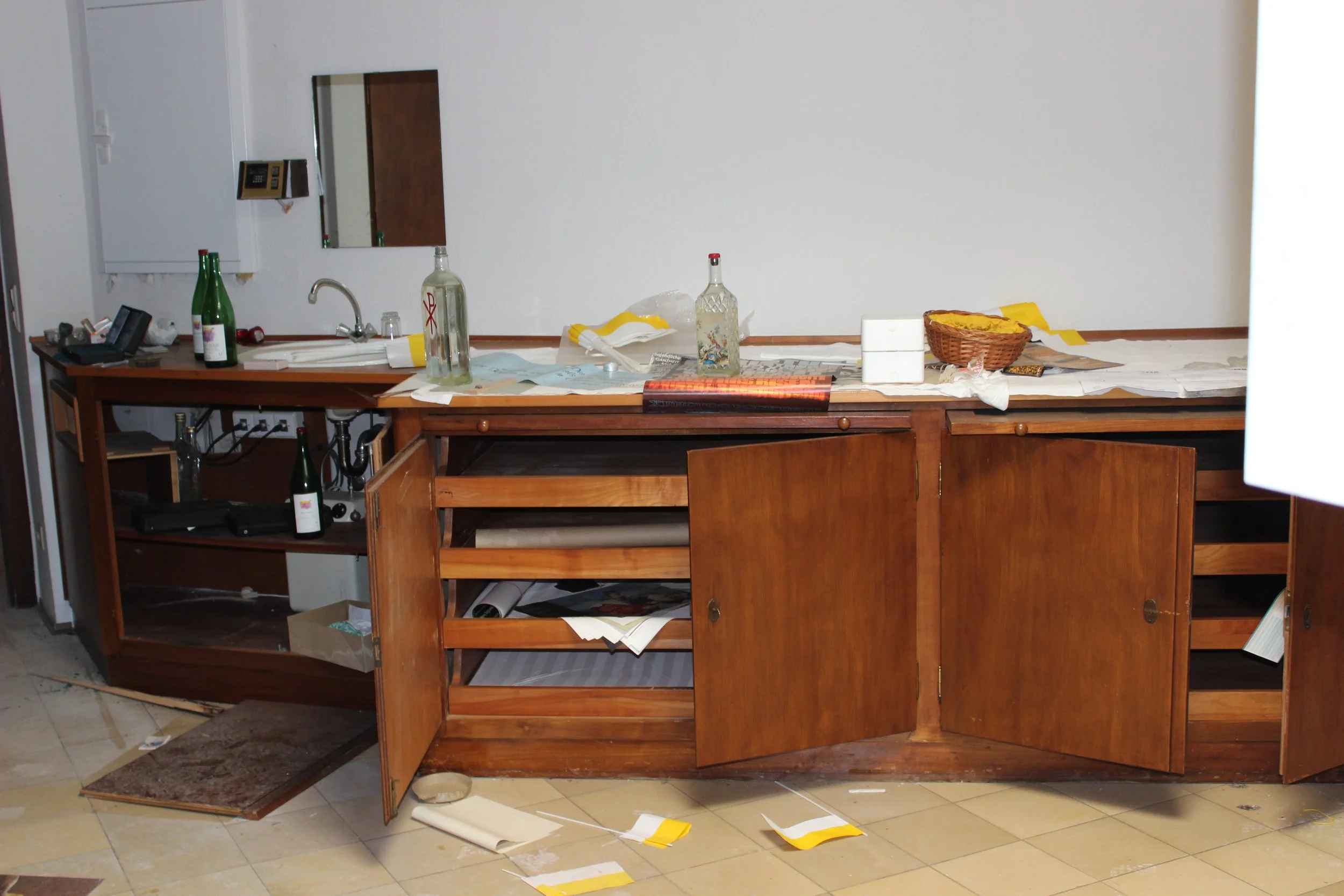 Unordentliche Küchenschrank mit offenstehenden Türen, zerstreute Gegenstände wie Flaschen, Papier und ein Korb auf der weißen Arbeitsplatte.