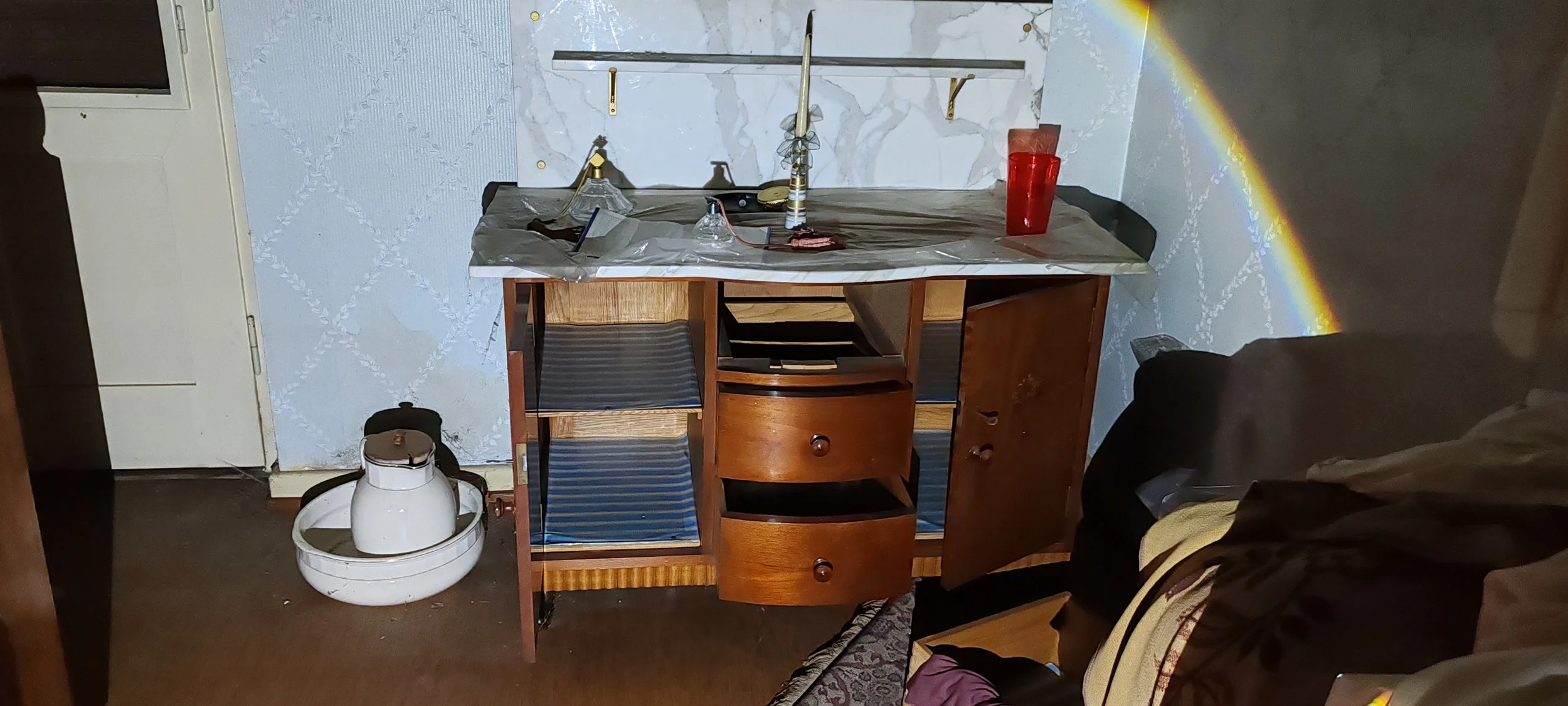 Küchenzeile mit Schrank, Wassertank, aufgeräumtem Tisch mit Wasserflasche und Gegenständen, und Regenbogenreflexion an der Wand.