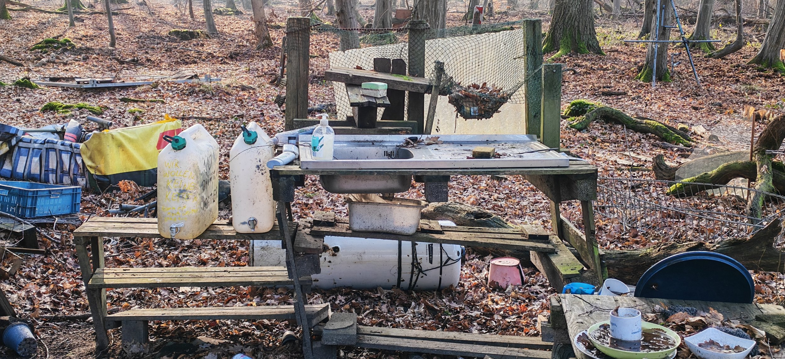 Verunreinigter Outdoor-Wasserbereich mit altem Waschbecken, gelben und weißen Kanistern, schmutzigen Geschirrresten, und Müll im Wald