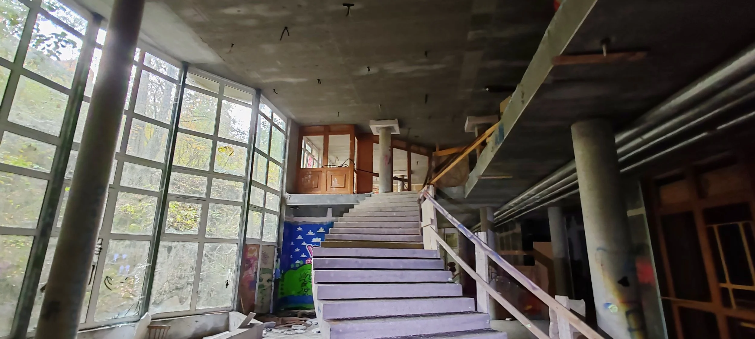 Innenaufnahme eines unvollendeten Gebäudes mit einer Treppe, großen Fenster, sichtbaren Rohren und Wandkunst.