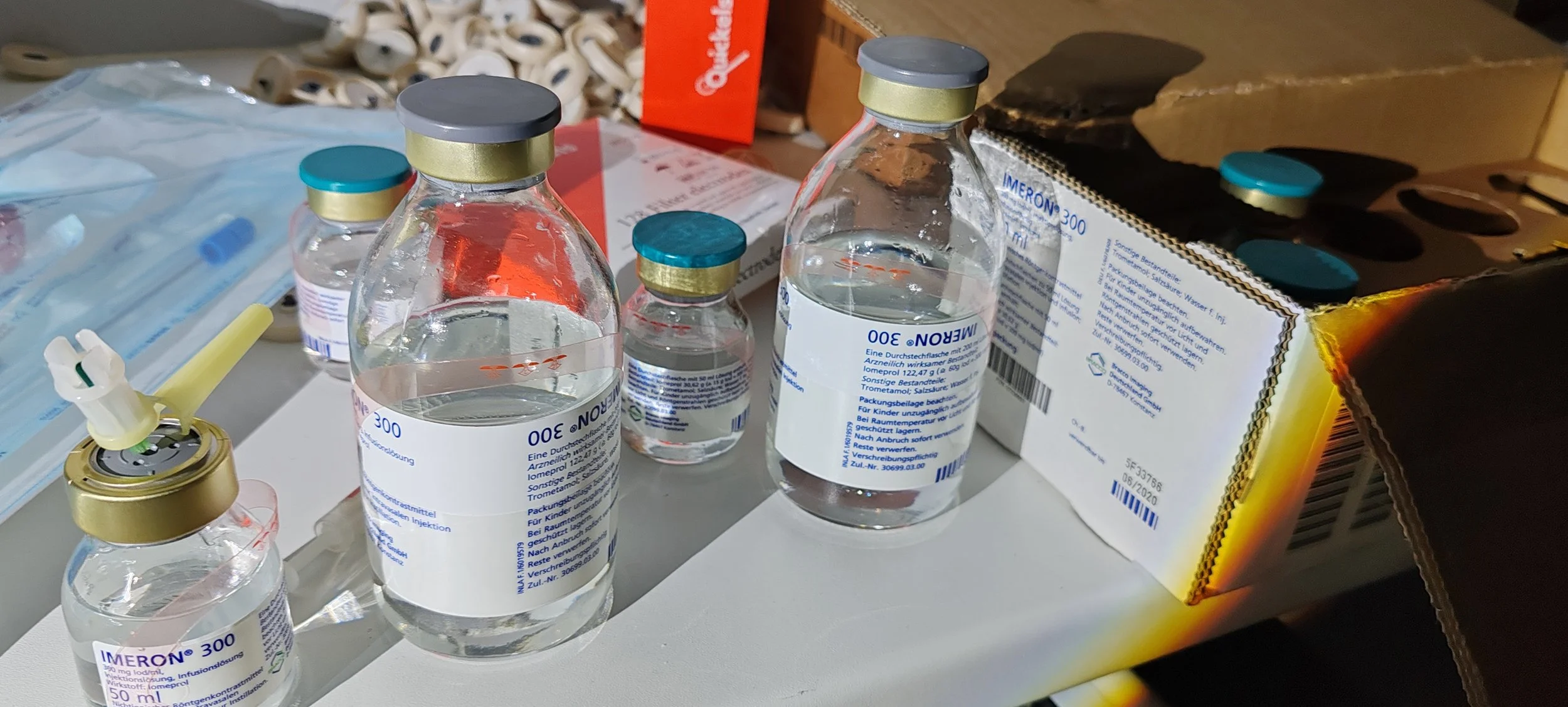 Verschiedene kleine Glasflaschen mit Medikamenten, darunter ein Impfstofffläschchen mit gelbem Verschluss, eine Kondensator-Infusionsflasche mit blauer Beschriftung, auf einem weißen Oberflächen mit einer stückweise geöffneten Medikamentenschachtel u