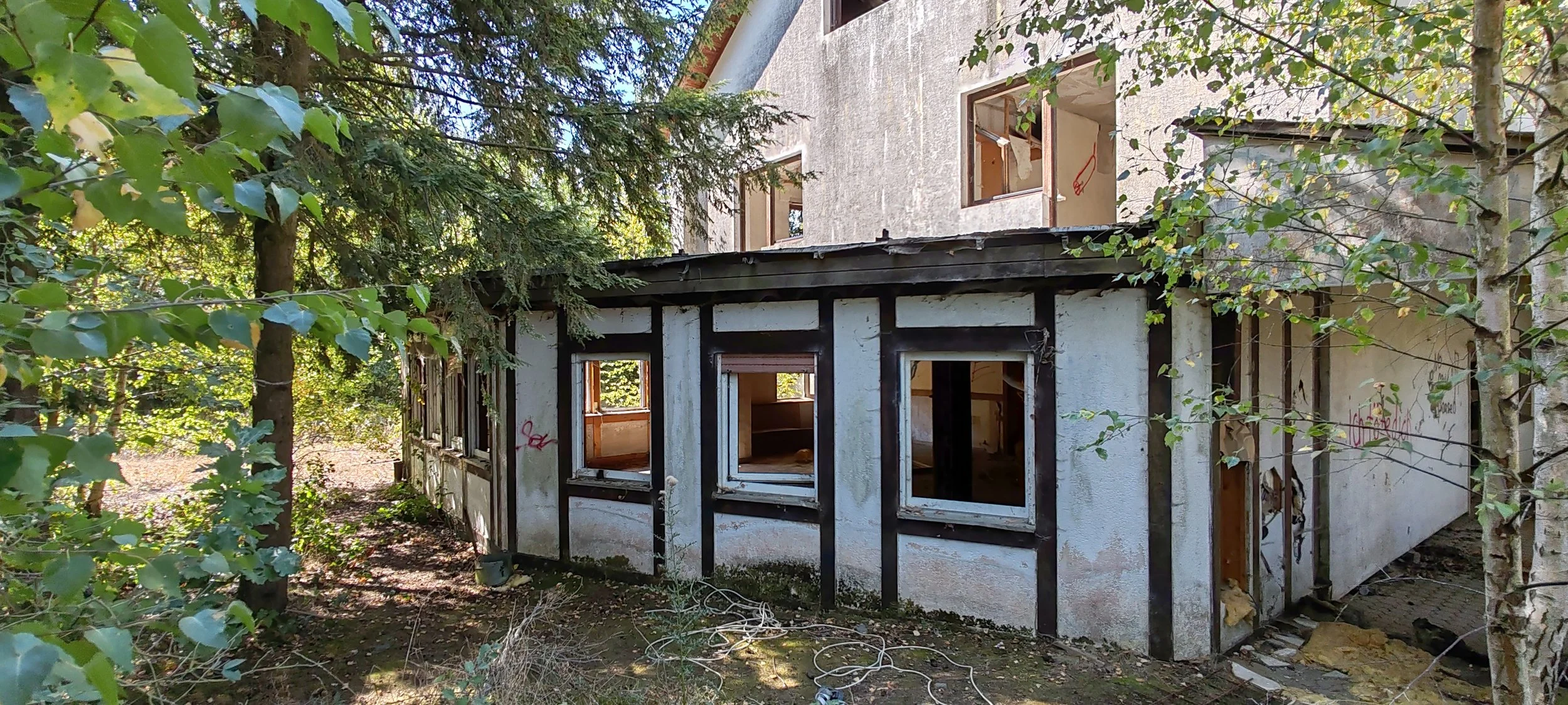 Das Bild zeigt ein altes, teilweise verfallendes Gebäude im Freien, umgeben von Bäumen und Vegetation. Das Gebäude hat Fensterrahmen ohne Fenster und ist im Bau oder Abrisszustand.