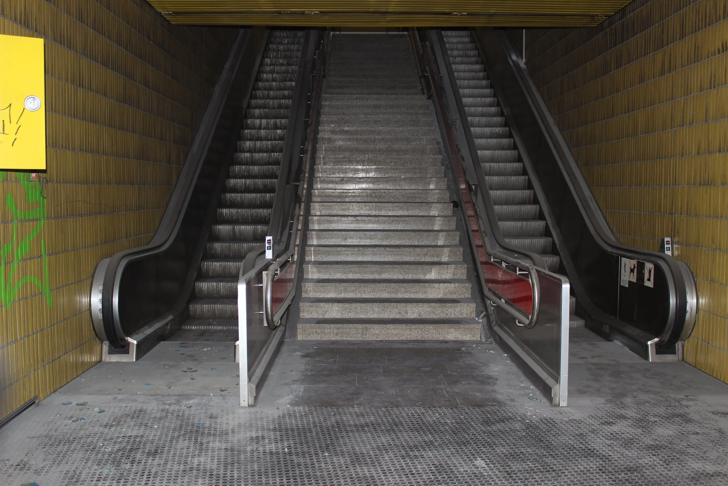 Ein Aufzug mit zwei Rolltreppen links und rechts, die in eine unterirdische Station führen, vor einer Wand mit gelben Fliesen.