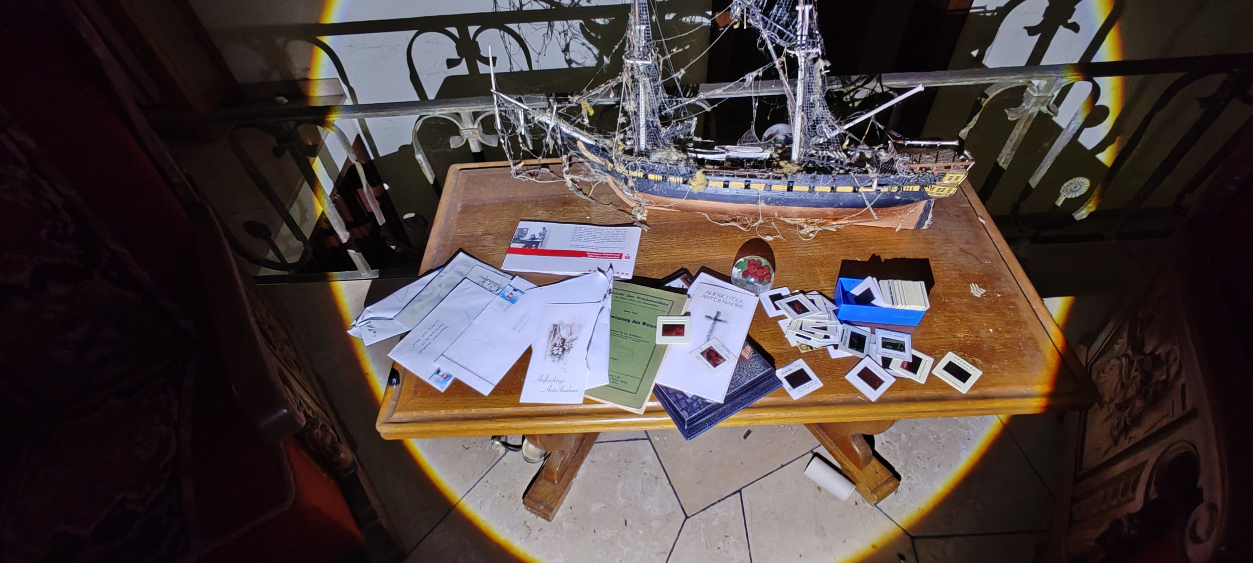 Ein Holztisch mit Papieren, Fotografien, einem Modell eines Segelschiffs und einer kleinen Dose mit roten Beeren. Im Hintergrund Schattenstrahlen sind auf die Wand projiziert.
