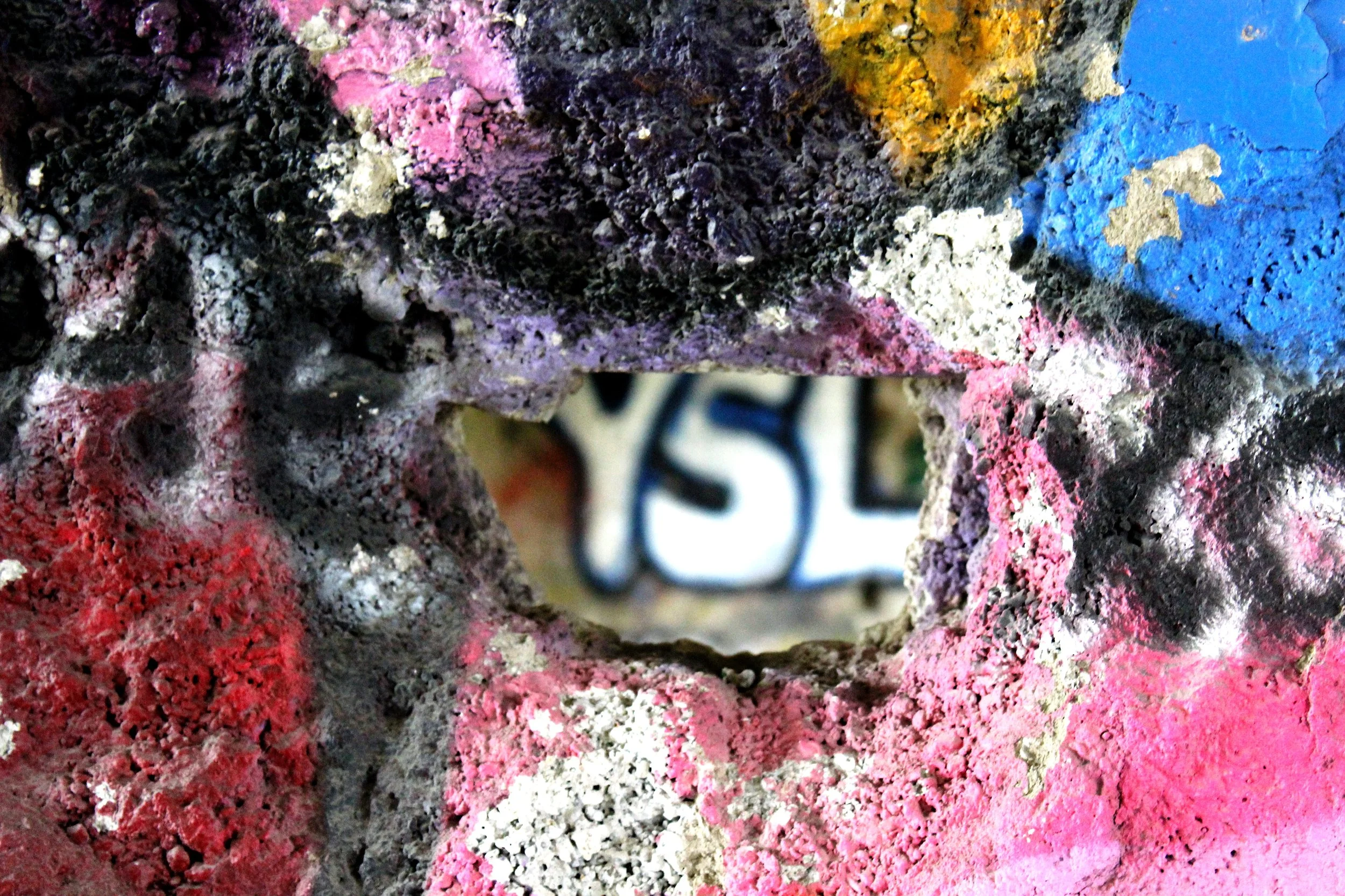 Blick durch ein Loch in einer bunten, verzierten Betonwand, mit dem Buchstaben 'YSL' erkennbar im Hintergrund.