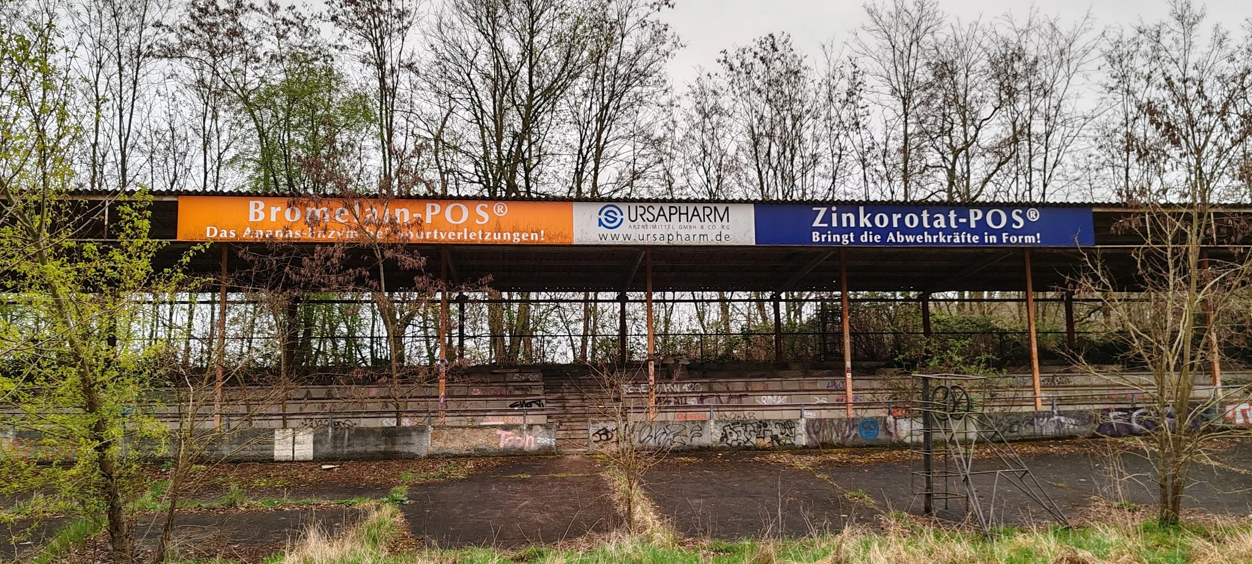 Leere, verlassene Tribüne im Freien, umgeben von Bäumen, mit Graffiti besprühter Wand und verrosteten Stufen, nachts oder bei trübem Wetter.