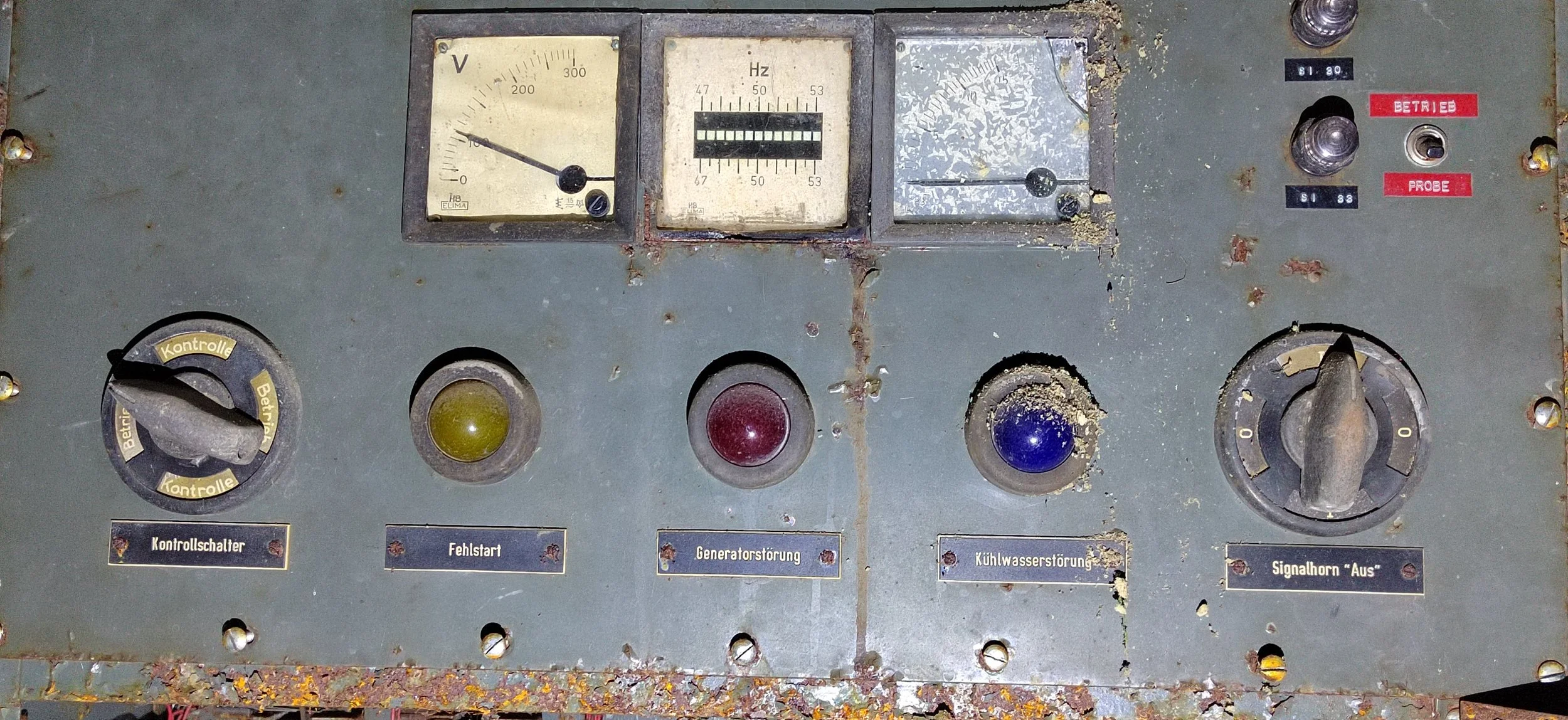 Altertes Kontrollpanel mit Schaltern, Ampeln und Messinstrumenten in einem industriellen Umfeld, beschriftet auf Deutsch.