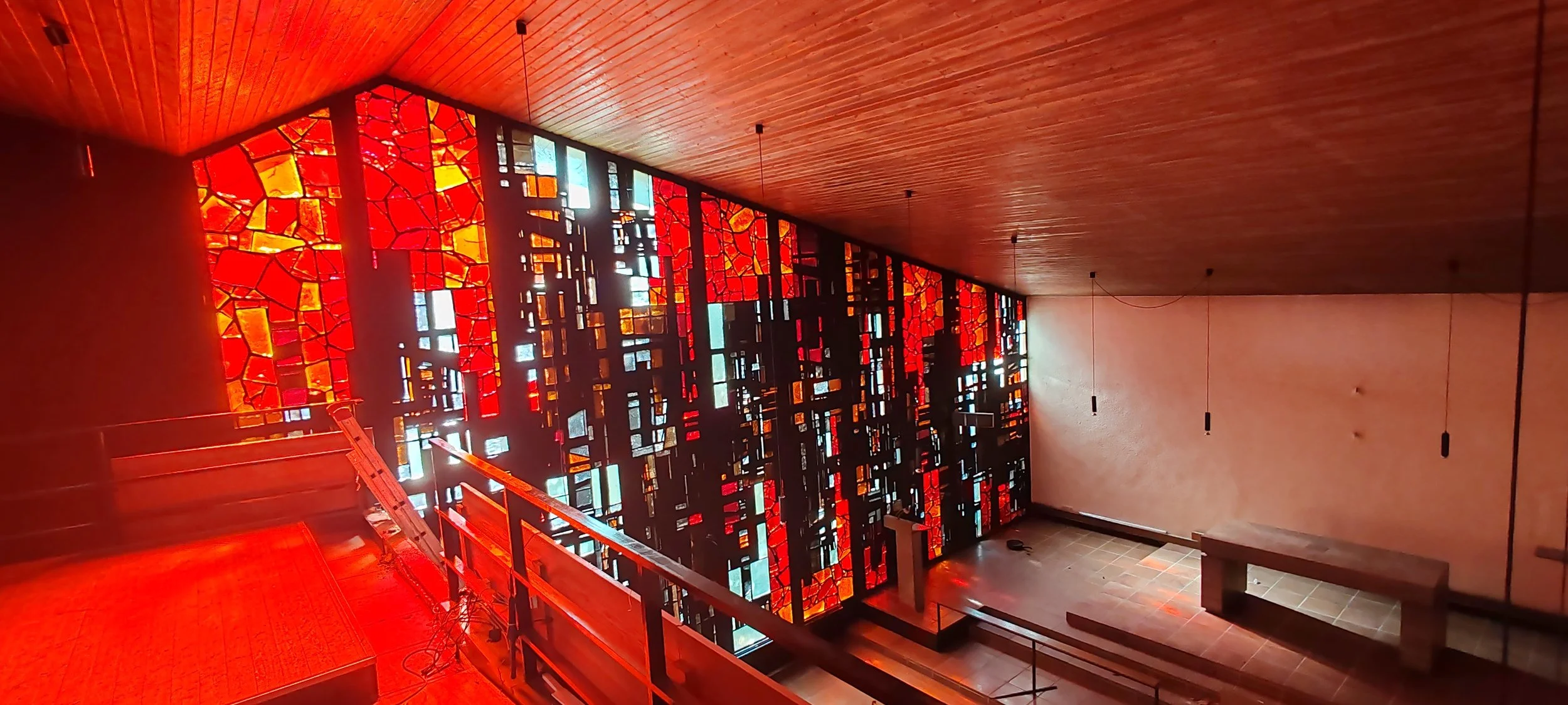 Innenraum eines modernen Gebäudes mit einer großen, farbigen Glasfensterwand in Rot- und Orangetönen und holzvertäfelter Decke.