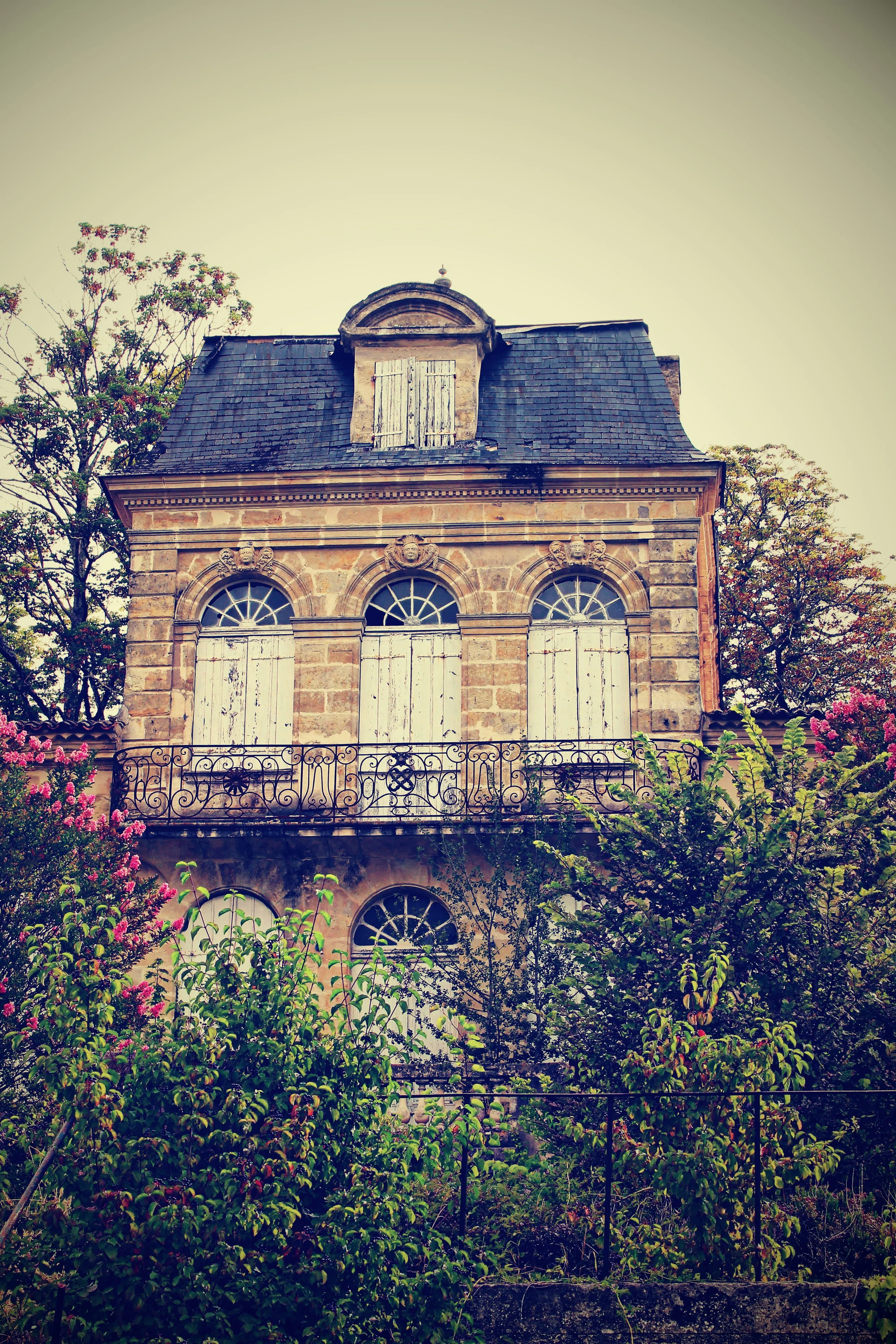 Ein altes, mehrstöckiges Haus mit steingemauerten Wänden, einem Schieferdach und geschlossenen weißen Fensterläden, umgeben von blühenden Büschen und Bäumen.