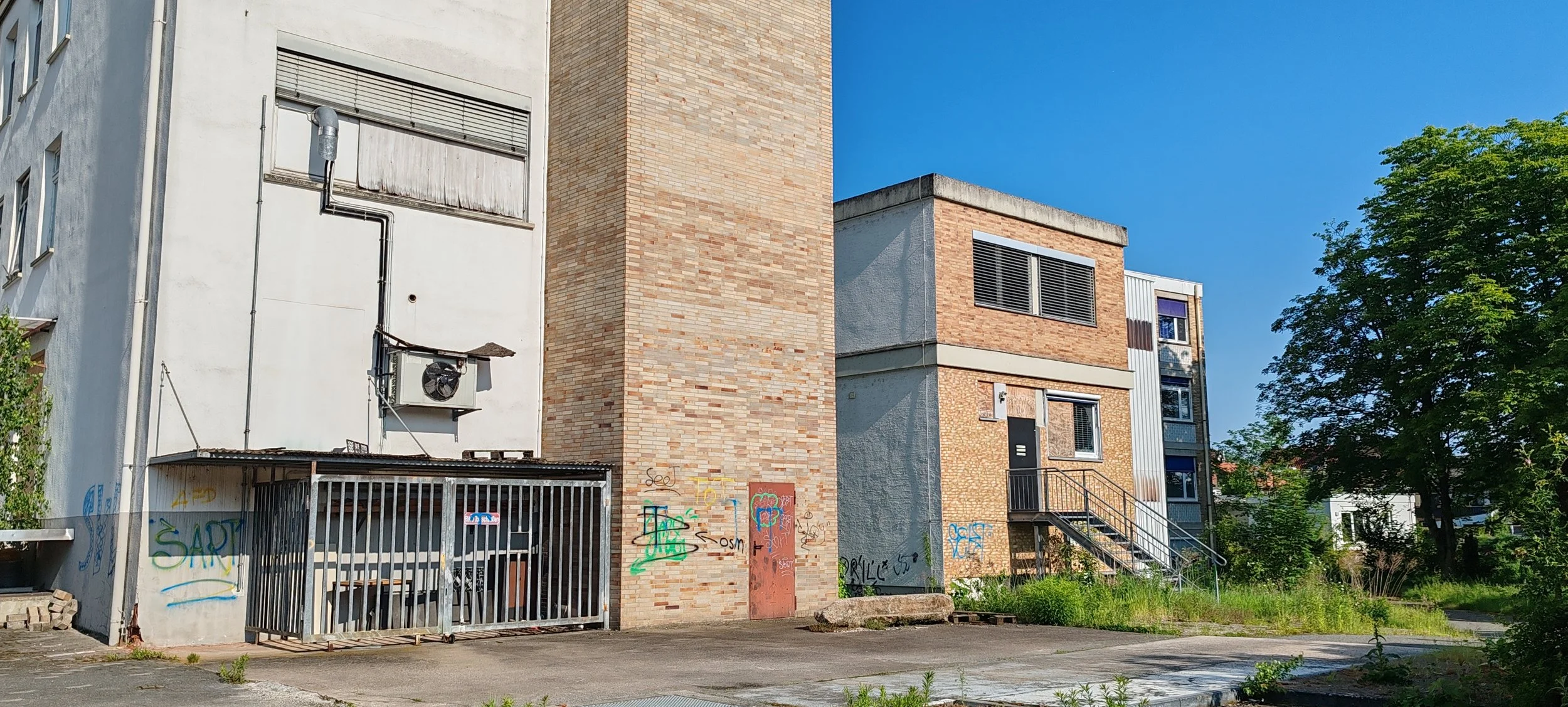 Mehrfamilienhaus mit Graffiti an der Wand, Brennholzstapel, Metalltreppe, grüner Baum im Hintergrund, blauer Himmel