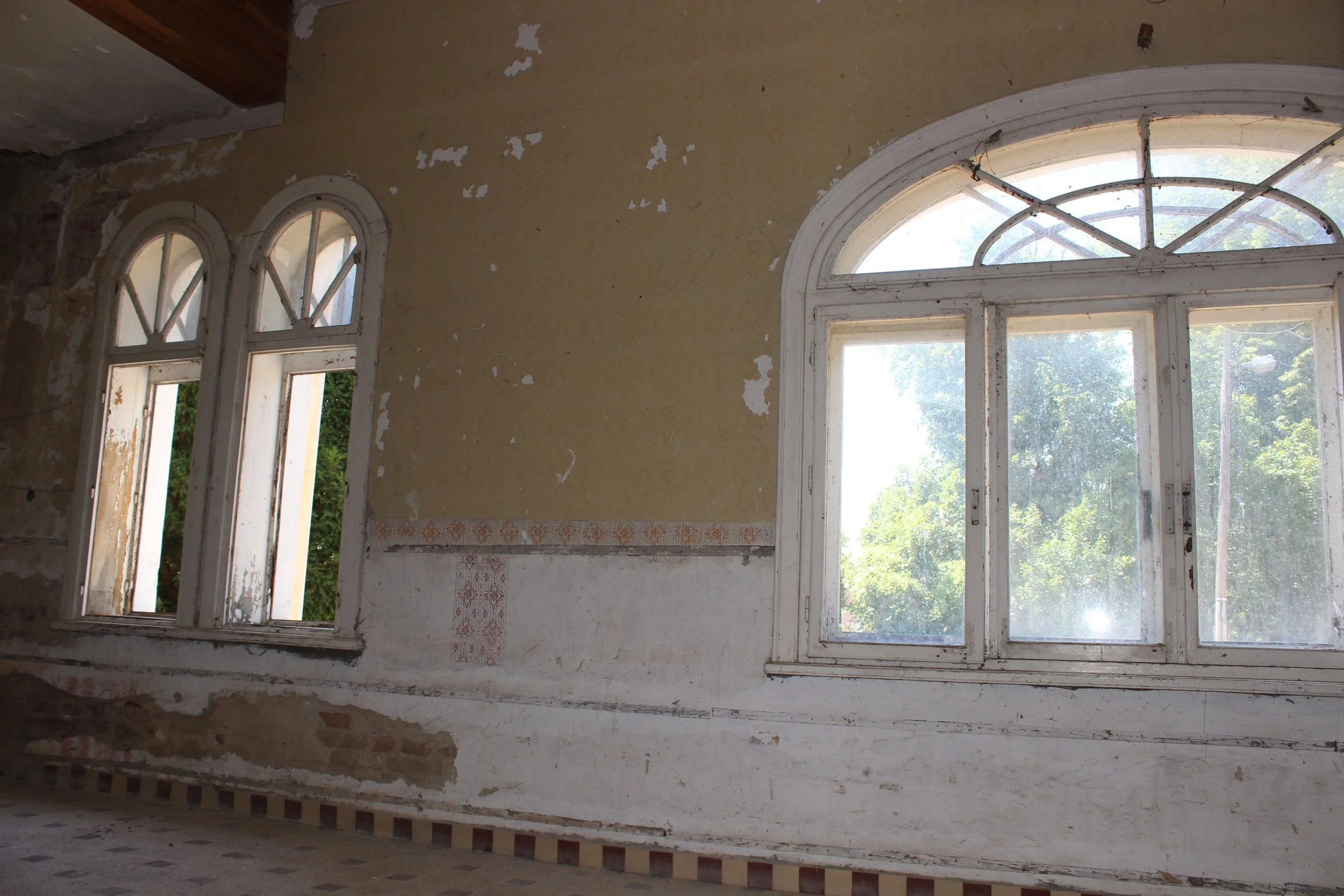 Innenansicht eines alten, verzierten Hauses mit großen, weißen, bogenförmigen Fenstern, die auf eine grüne, helle Umgebung blicken. Die Wände sind teils abblätternd und teilweise verputzt, mit sichtbaren Spuren von Renovierungsarbeiten.