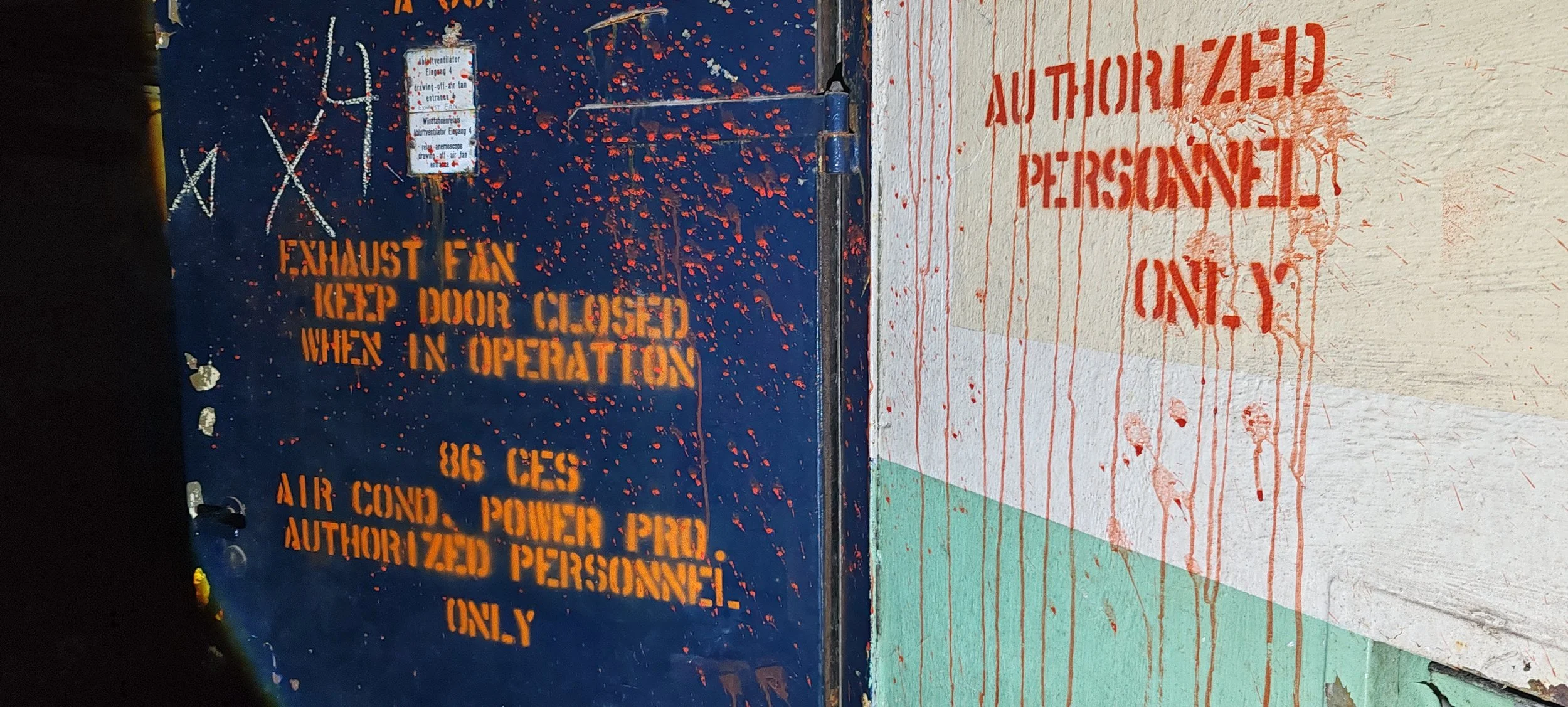 Warnhinweis in englischer und deutscher Sprache auf einer Tür, dass das Explosivfeld bei Betrieb geschlossen bleiben muss. Die Tür ist dunkelblau mit roten Farbspritzern, darüber liegt ein kleiner Zettel.