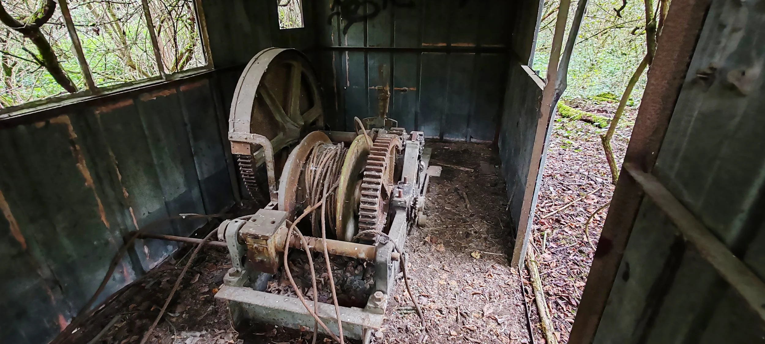 Alter Maschinenraum, rostige technische Geräte mit Zahnrädern in einer verlassenen Hütte im Wald, umgeben von Bäumen und Natur.