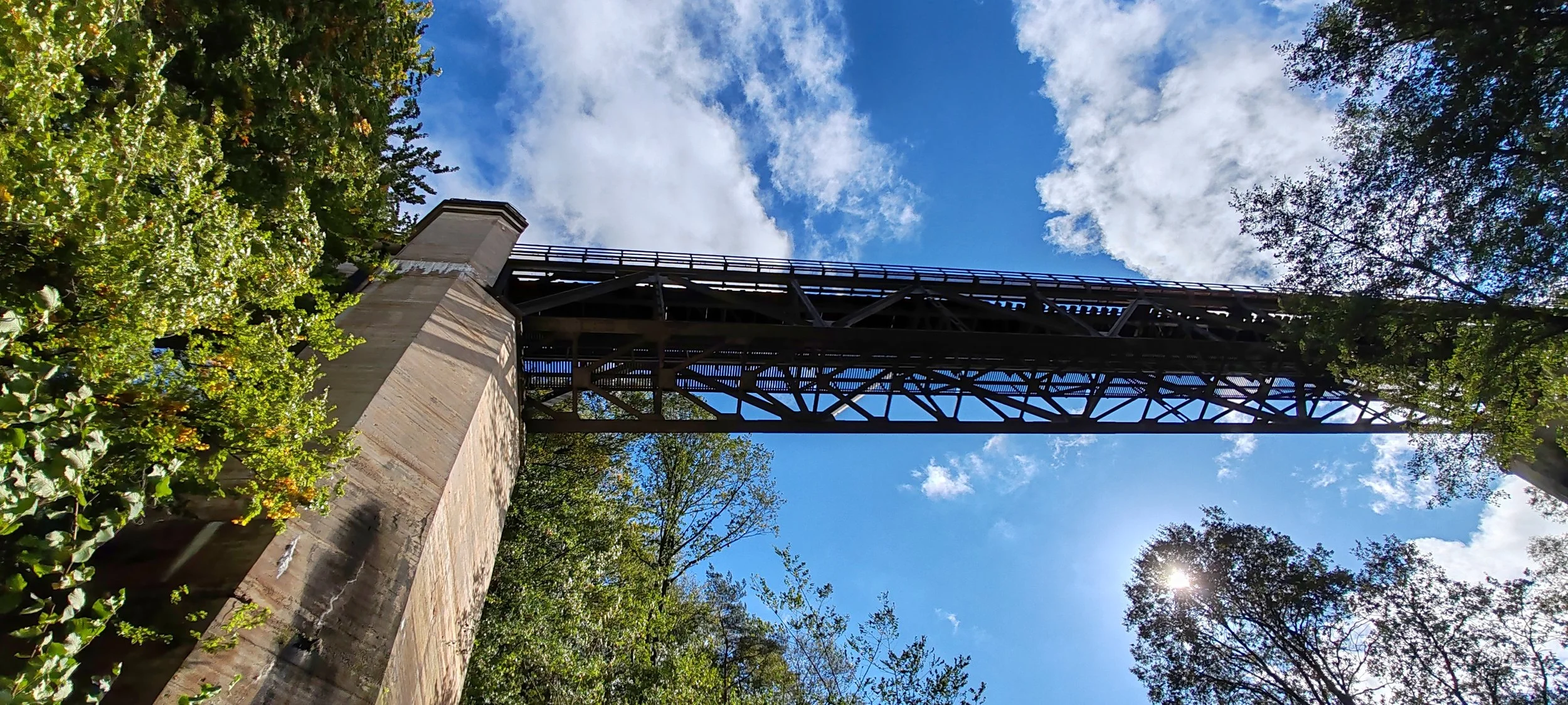 Blick nach oben auf eine Brücke, umgeben von Bäumen bei sonnigem Wetter und blauen Himmel mit Wolken.