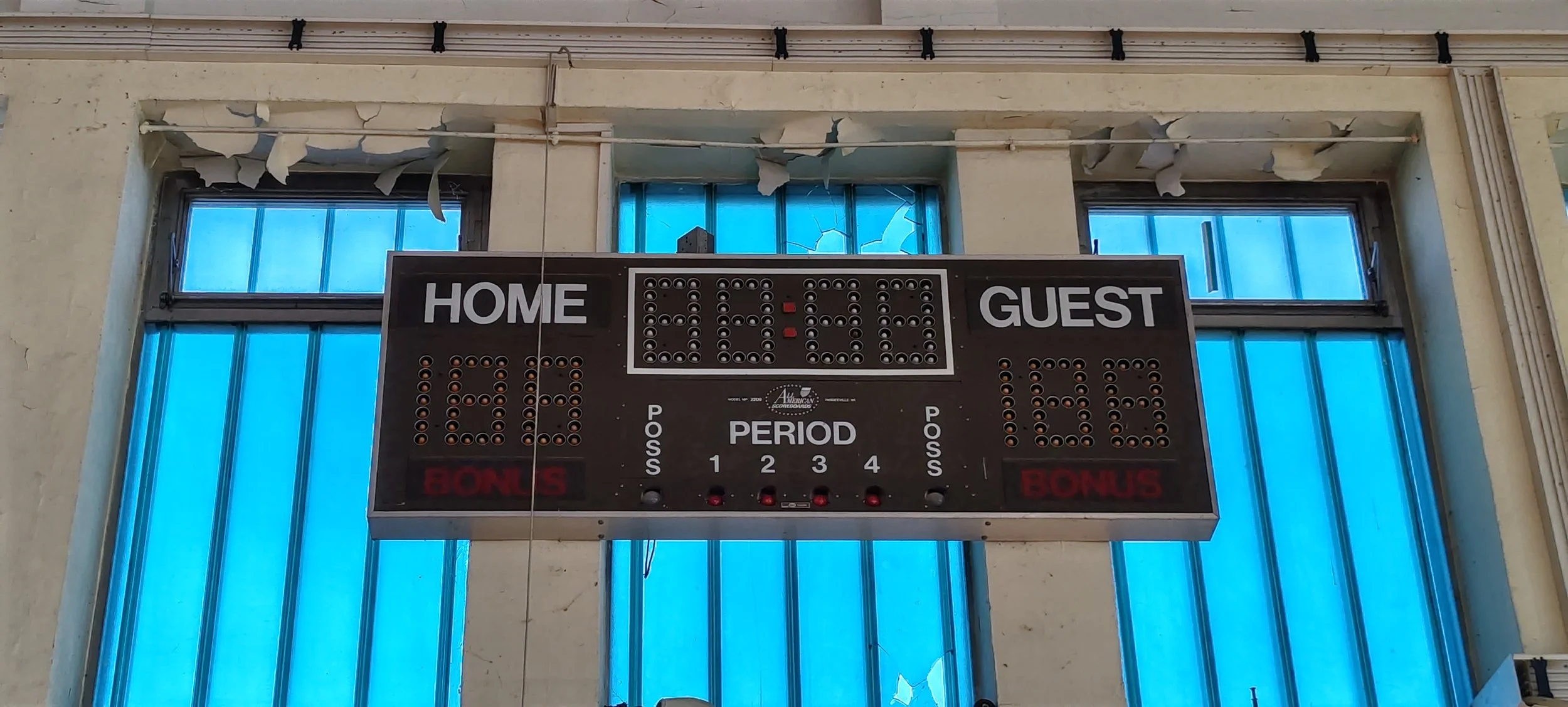 Ein Turnierraster für eine Basketballuhr, das an einer defekten Wand hängt, mit der Beschriftung 'Home', 'Guest', und einer digitalen Anzeige. Die Wand dahinter ist beschädigt, mit abblätternder Farbe und zerbrochenen Fenstern über dem Raster.