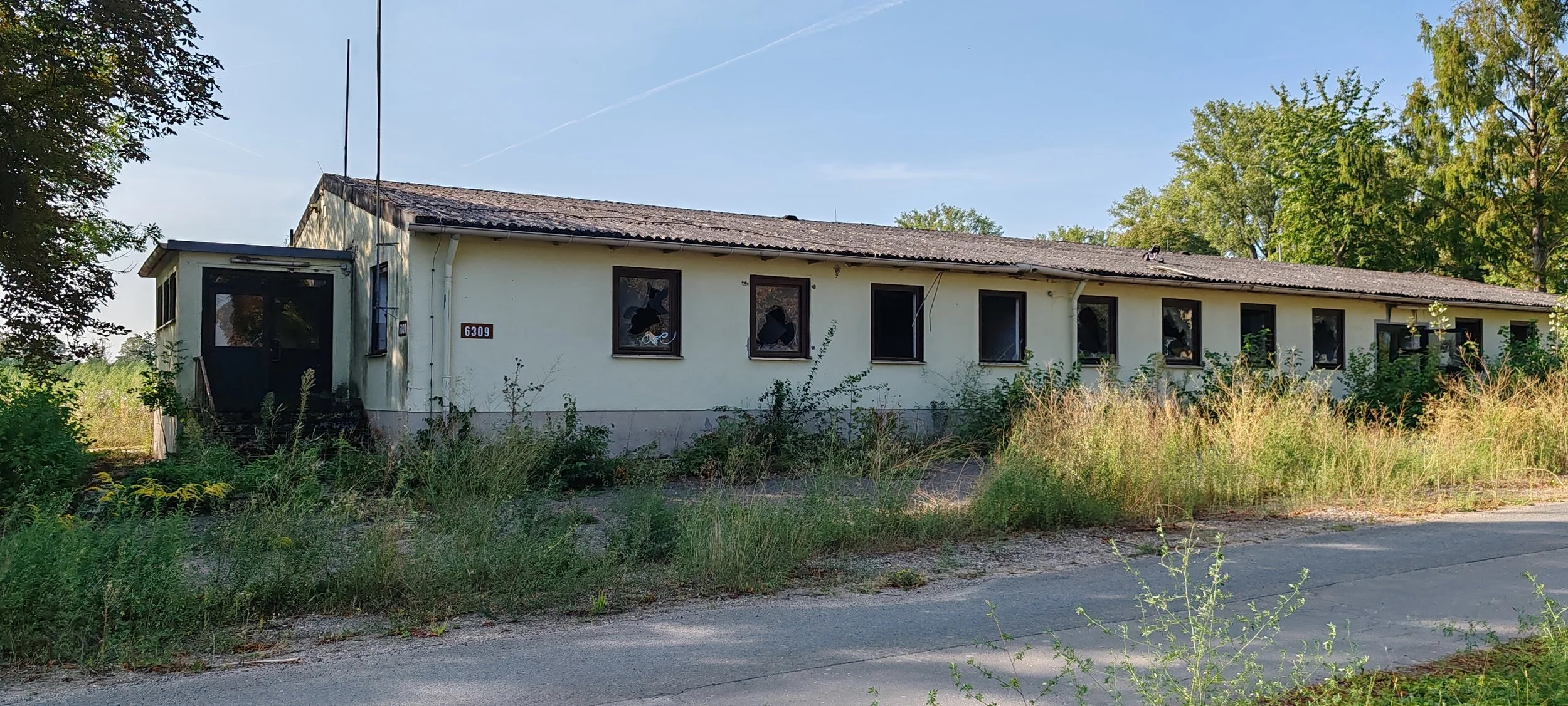 Ein verlassenes, zerfallenes altes Gebäude mit beschädigten Fenstern, umgeben von überwucherter Vegetation und Wäldern im Hintergrund.