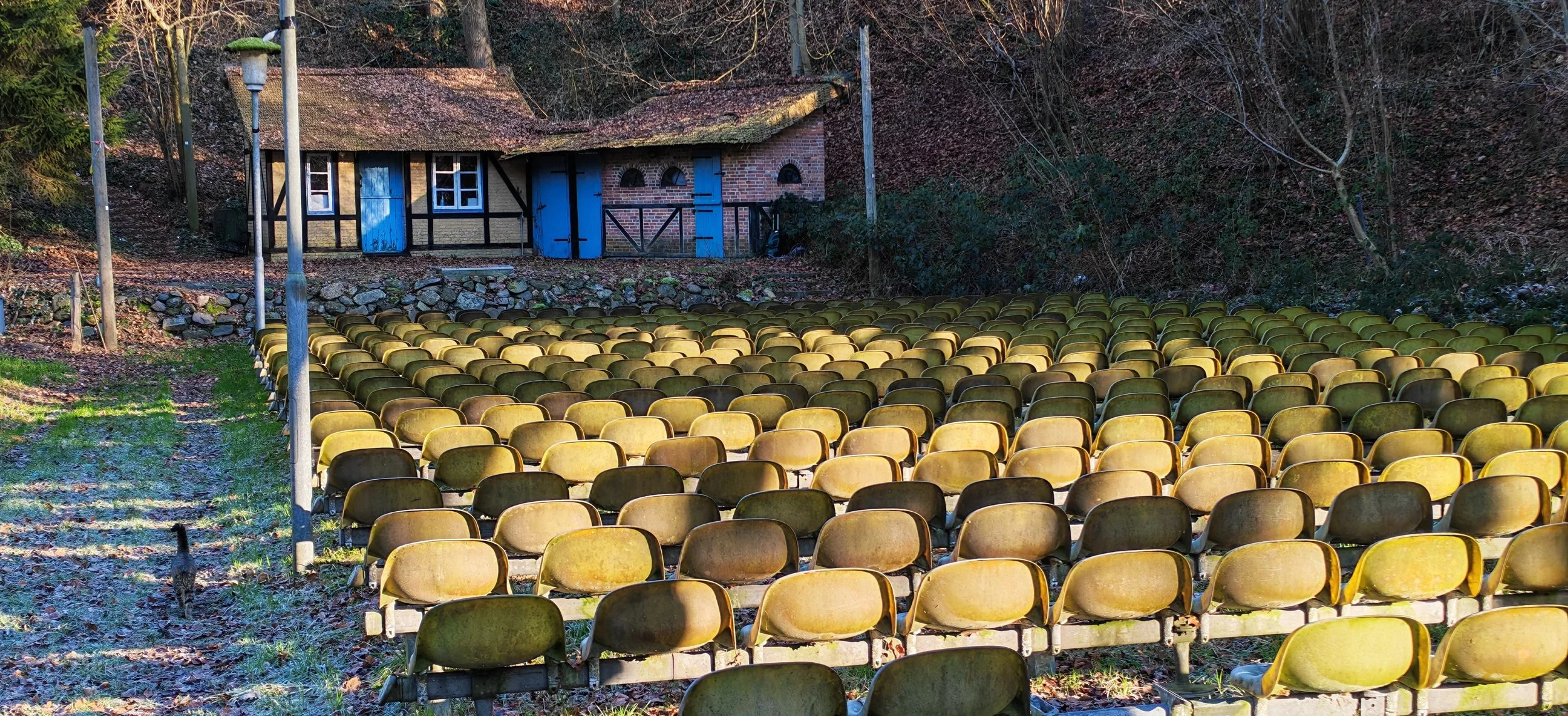 Leere, leicht verfallene Sitzreihen im Freien vor einem kleinen, bunten Haus mit blauem Tür und Fenstern, umgeben von Laub und Bäumen im Herbst.