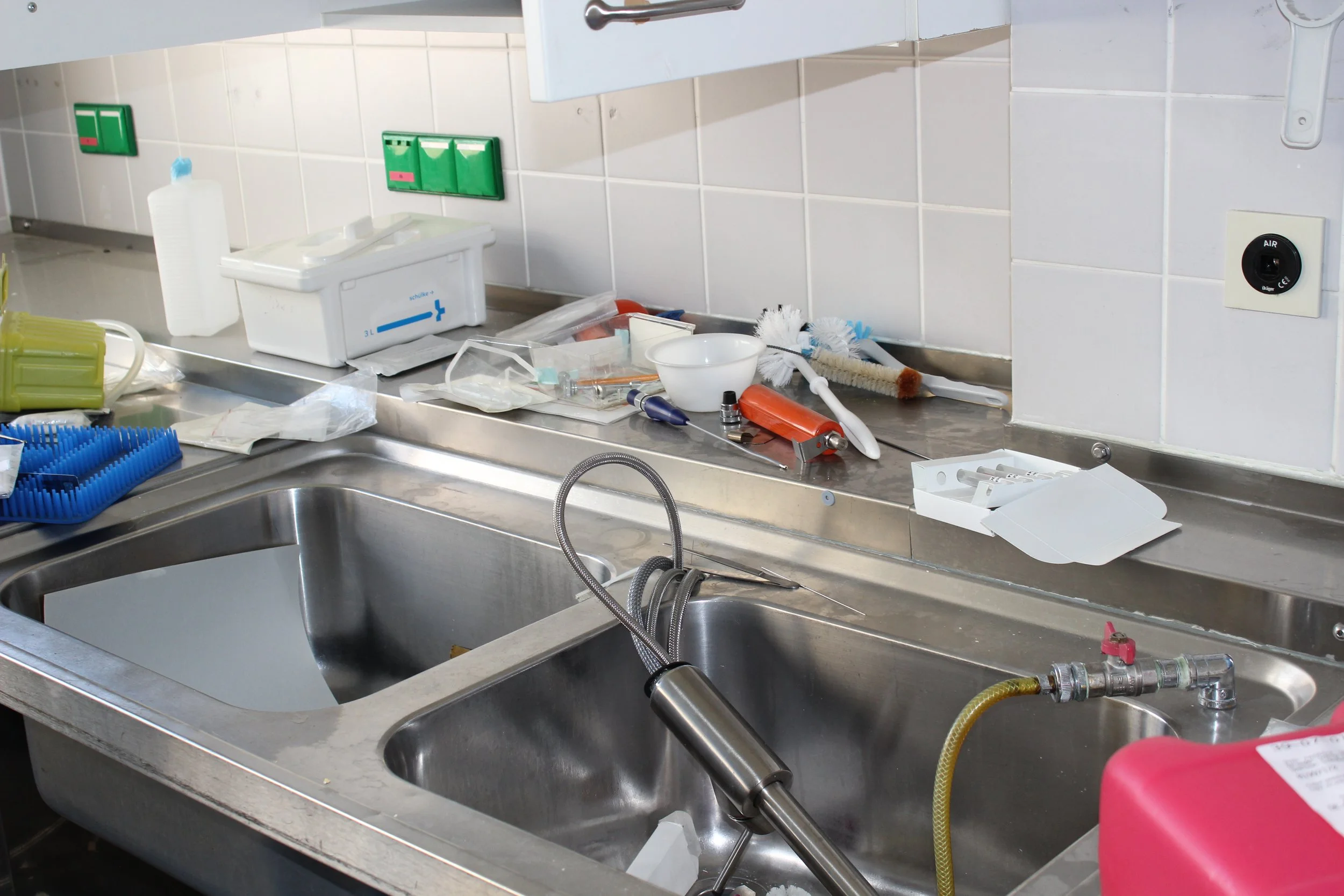 Unordentlicher Arbeitsbereich in einer medizinischen oder wissenschaftlichen Einrichtung mit Reinigungsgeräten, Spritzen, Reinigungsbürsten und anderen Werkzeugen auf der Edelstahl-Arbeitsfläche und im Spülbecken.