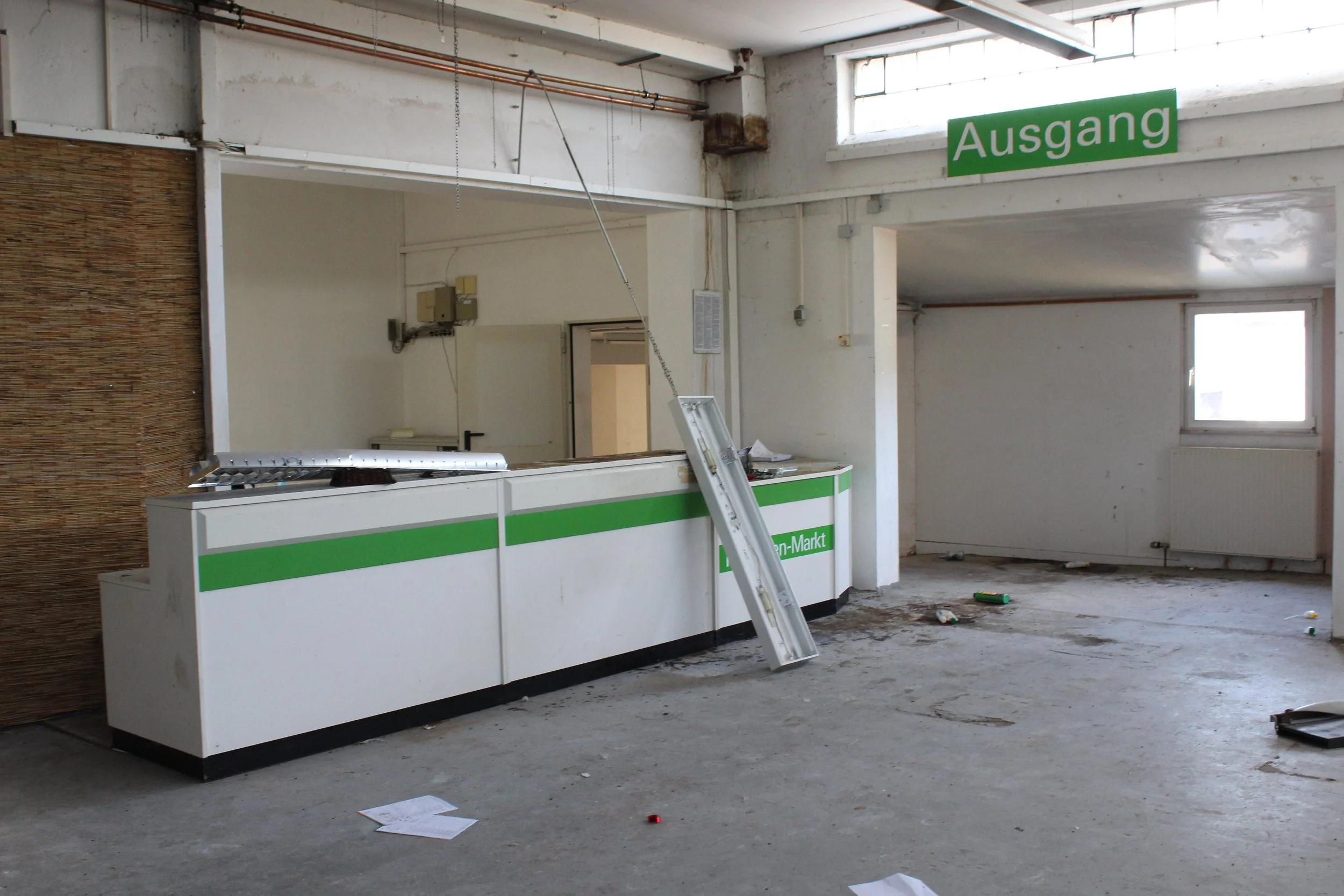 Leere Supermarkt-Kasse in einem verlassenen und ungepflegten Laden mit Papierkram auf dem Boden, einem umgestürzten Regal, einem offenen Stromkasten und einem grünen Schild mit der Aufschrift 'Ausgang'.