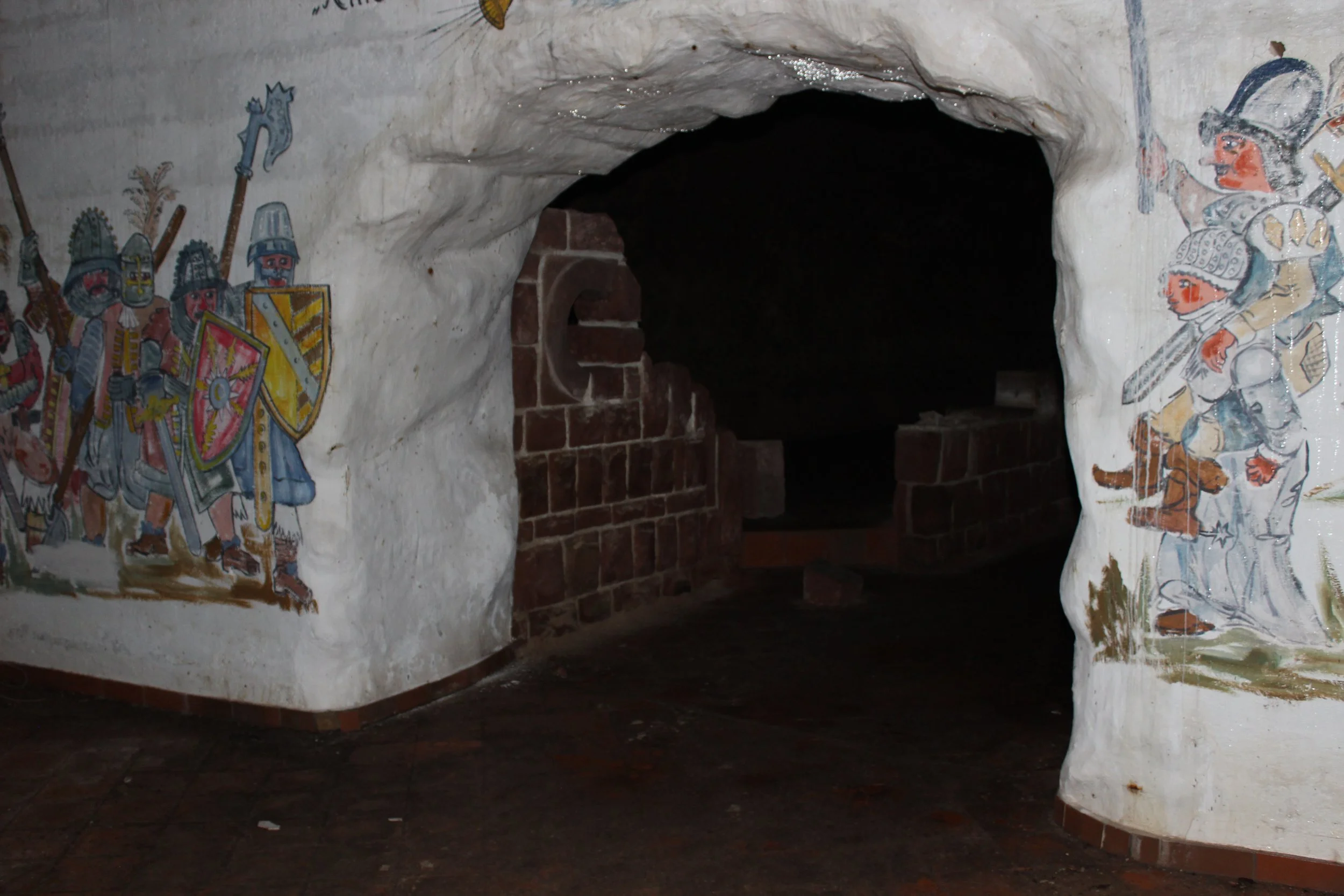 Unterirdischer Gang mit bemalten Wänden, die Ritter in Rüstung und mit Schilden zeigen.