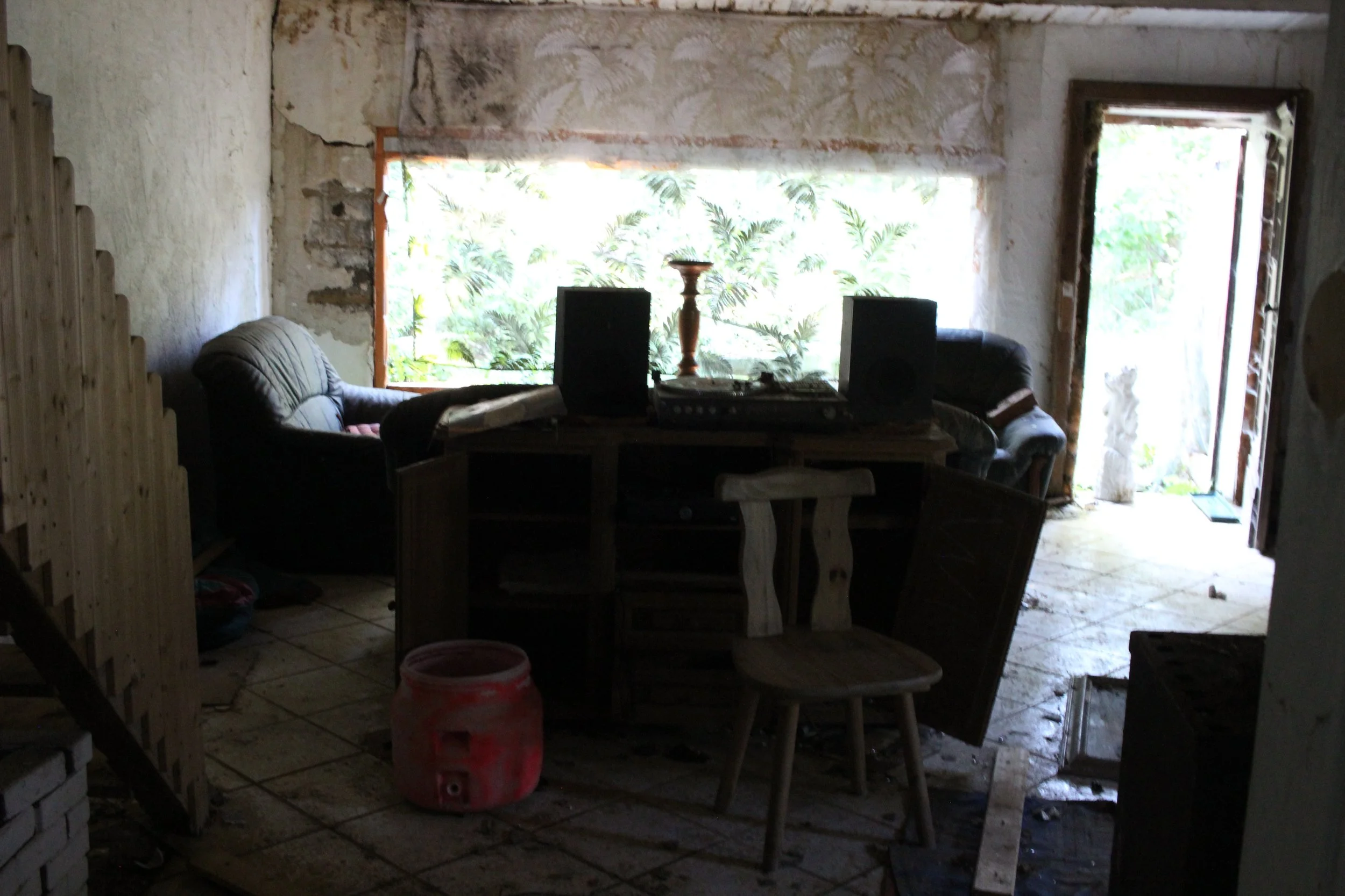 Ein dunkler, unordentlicher Raum mit zerbröckelten Wänden, einem Tisch mit Musik-Equipment, zwei Sofas, einem Stuhl und einem Krug auf dem Boden. Im Hintergrund sind große Fenster mit Blick auf grünes Außenland.