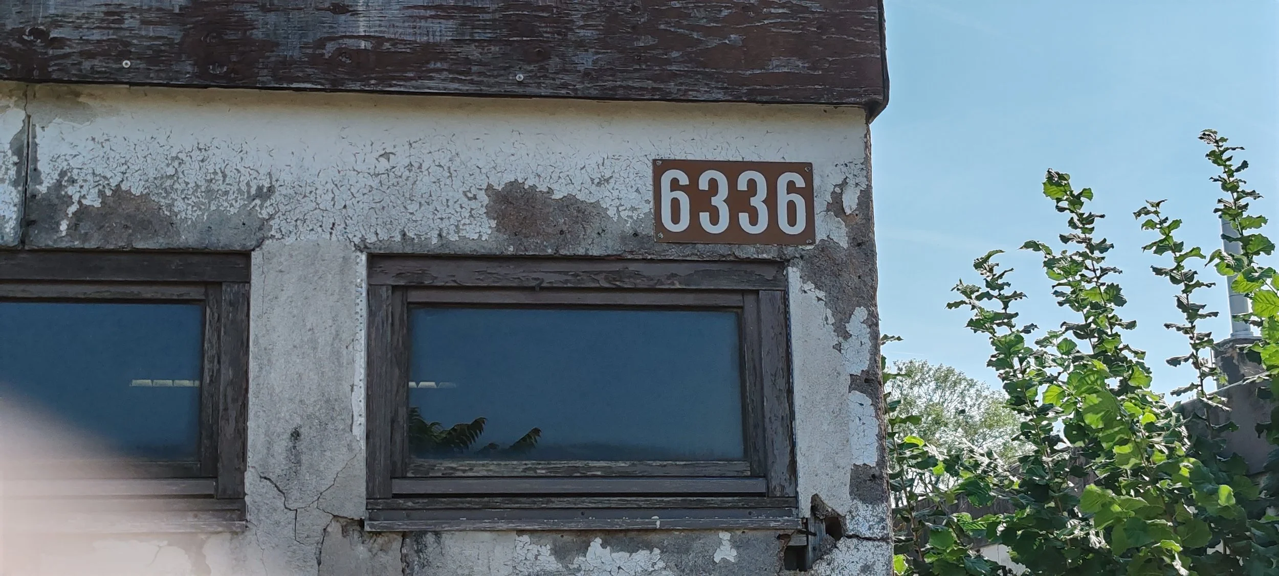 Zweistöckiges Haus mit braunen Holzelementen, zwei Fenster, eine Hausnummer 6336 und eine Pflanze mit grünen Blättern auf der rechten Seite.