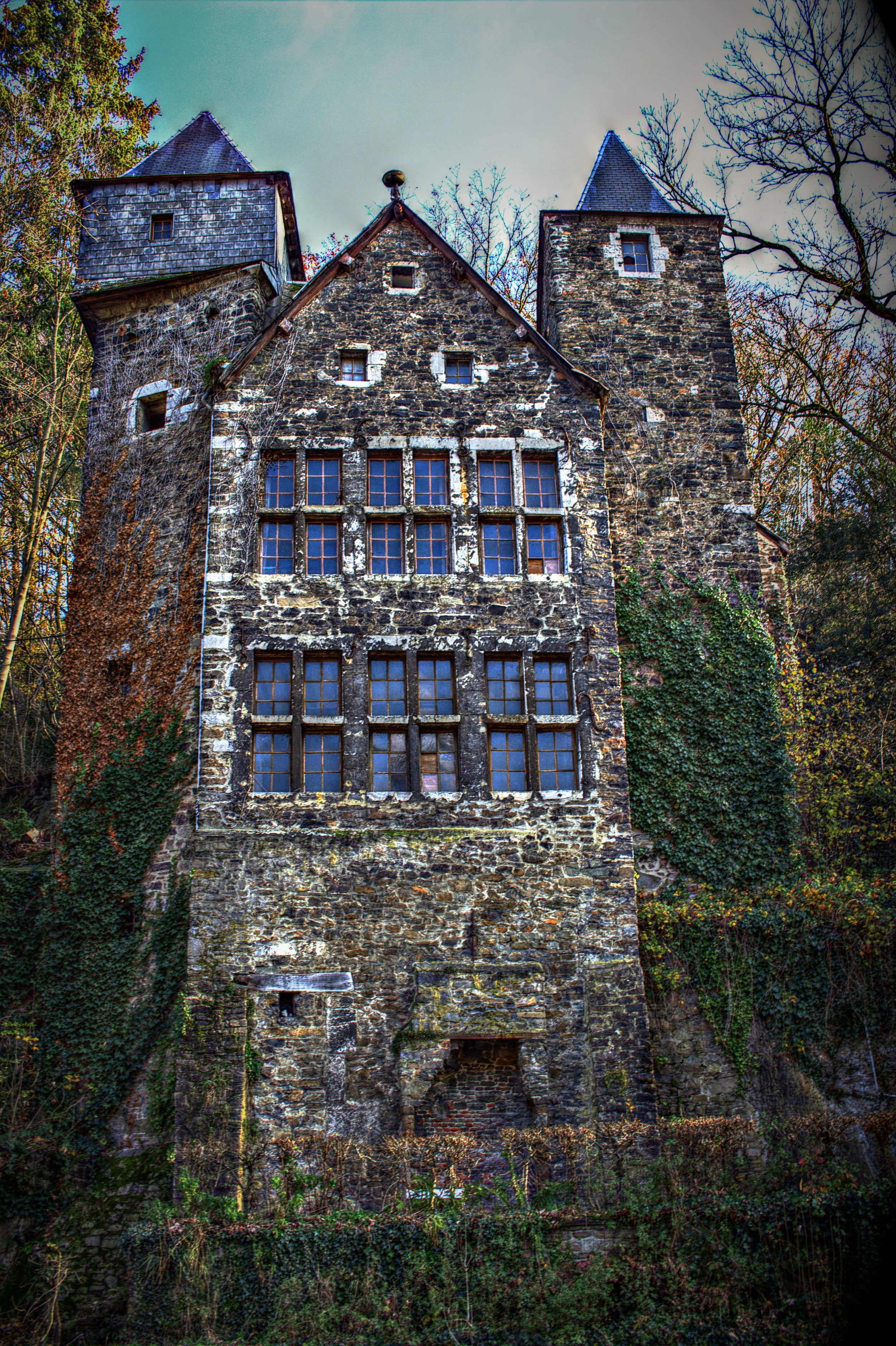 Ein altes, verfallenes Steingebäude mit mehreren Fenstern, umgeben von Bäumen und Efeu.