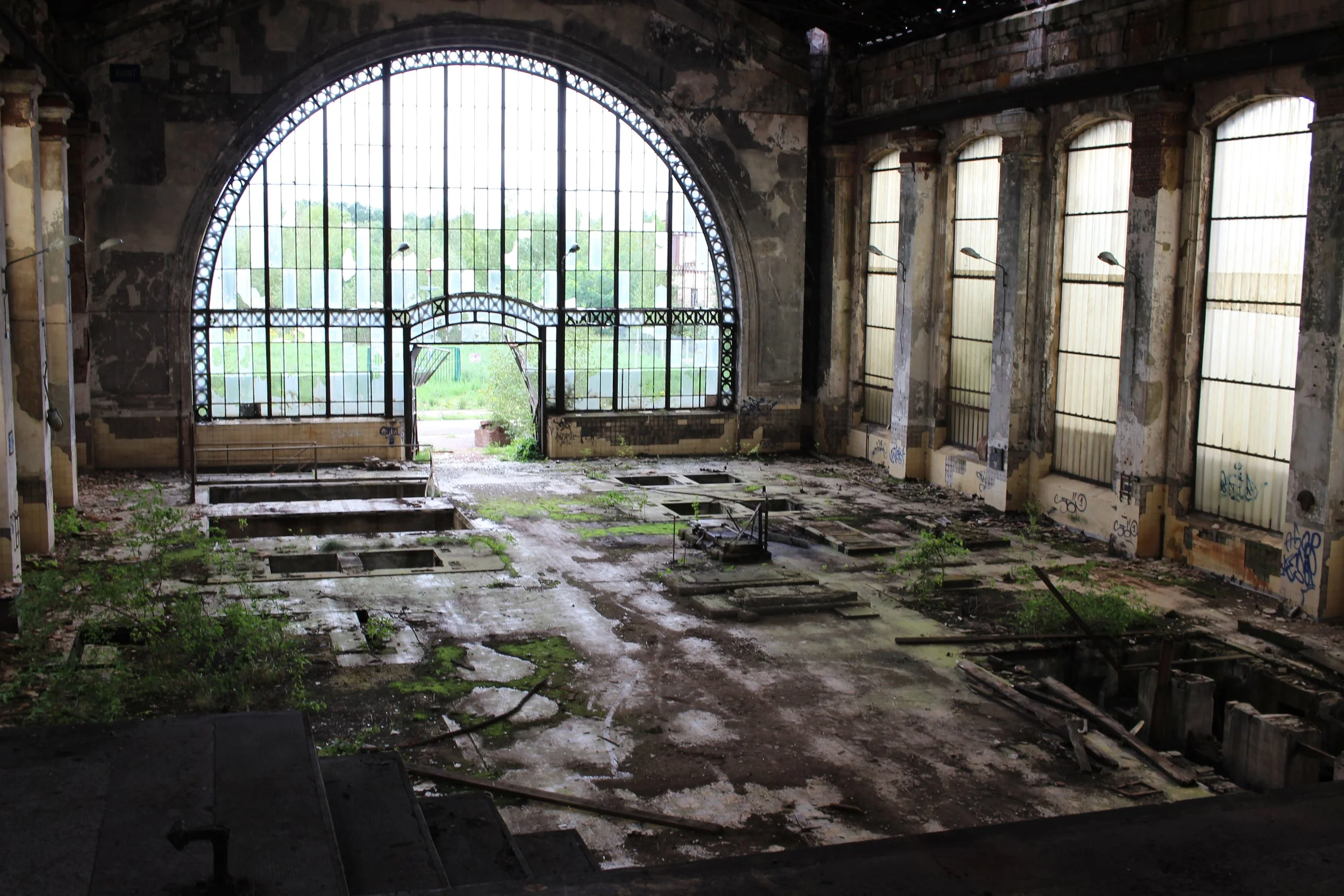 Verlassener, verfallener Saal mit großen Fenstern, Graffiti an den Wänden, dreckigem Boden und überwucherten Pflanzen