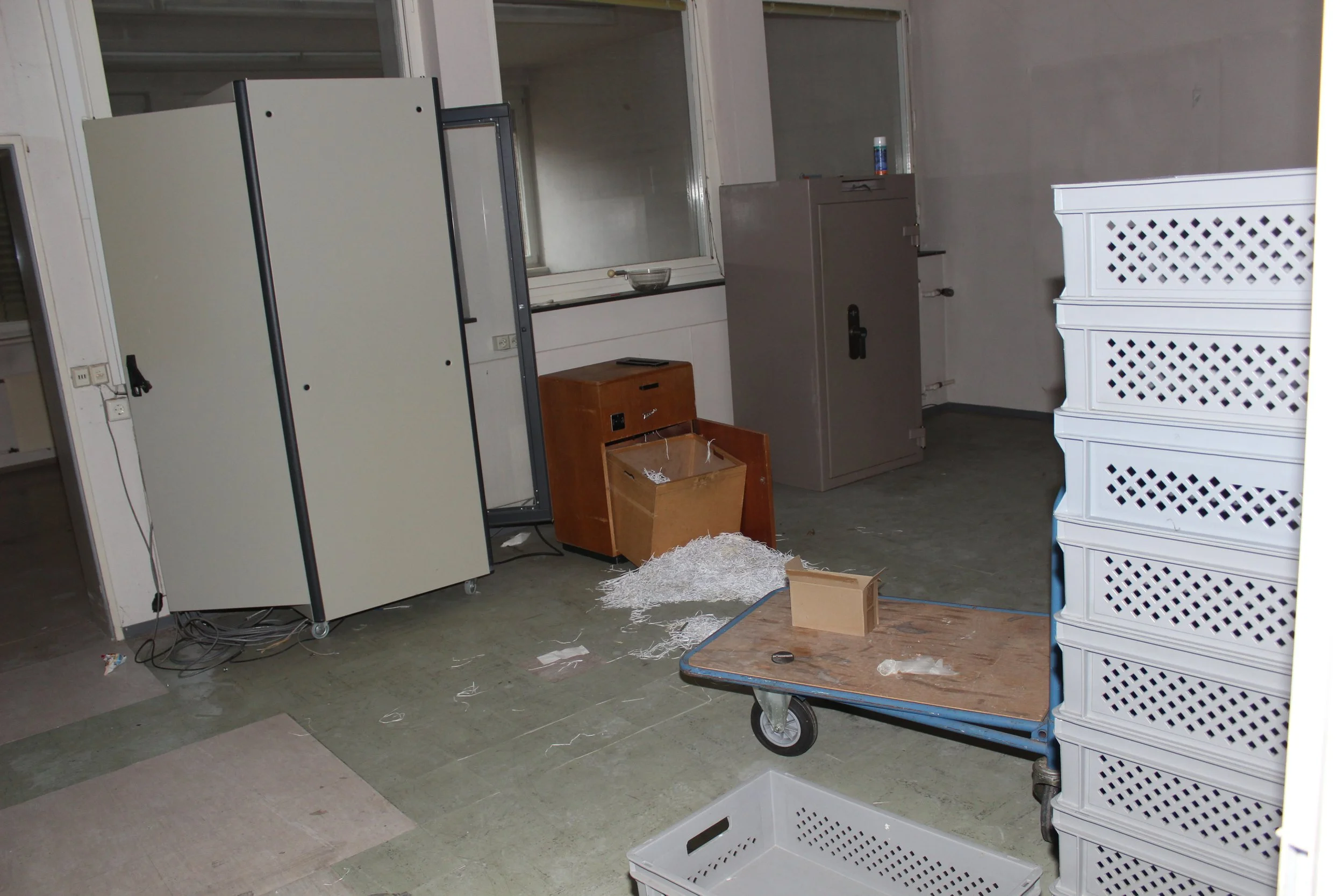 Unordentlicher Raum mit einer umgestürzten blauen Einkaufskarre, weißen gestapelten Kästen, einer braunen Schublade, einer Mülltonne und einer Metallablage, die überall verteilt sind.