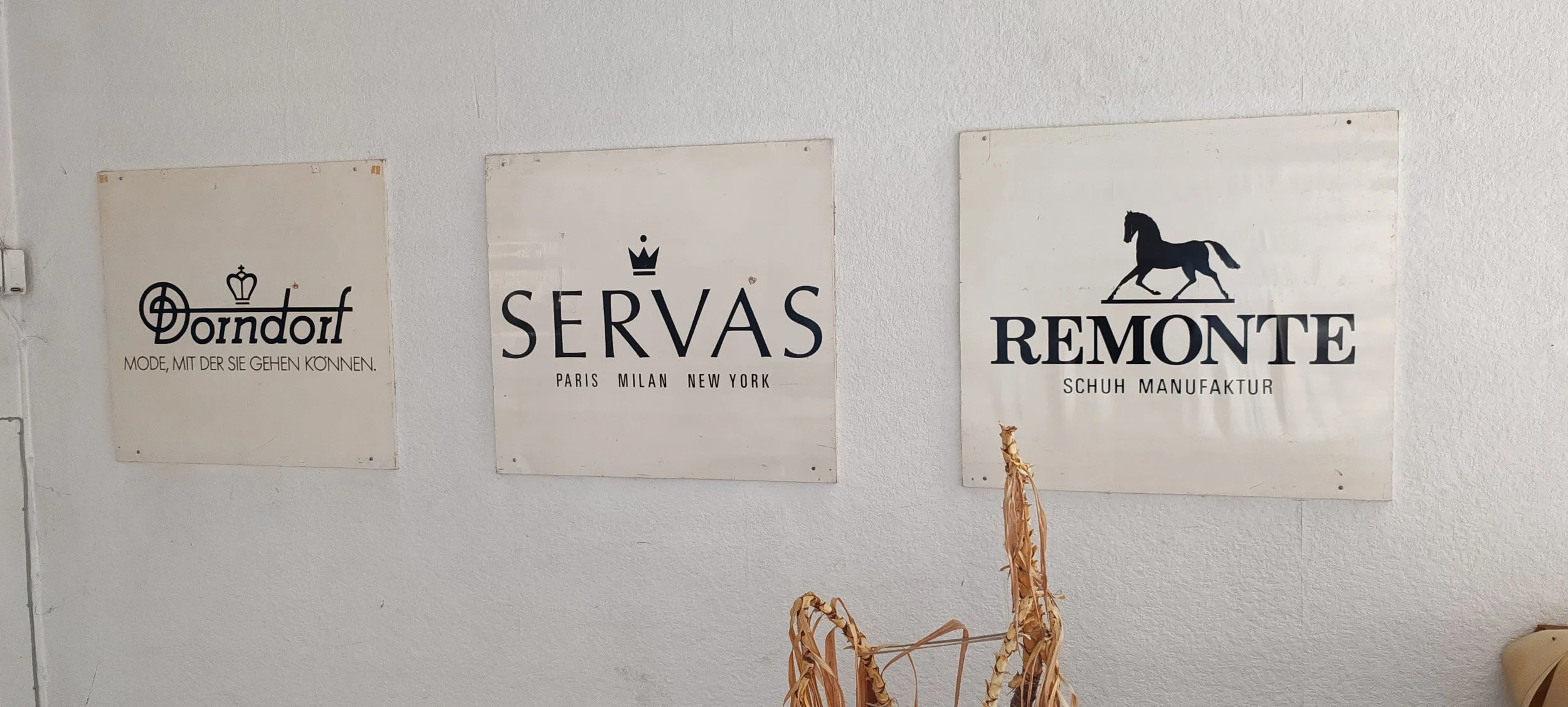 Drei weiße Werbetafeln mit schwarzen Logos und Texten, die an einer weißen Wand hängen. Darauf sind das Logo von Doendorf mit einem Krone-Symbol und der Slogan "Mode, mit der sie gehen können", das SERVÀS-Logo mit einer Krone und den Städten Paris, M