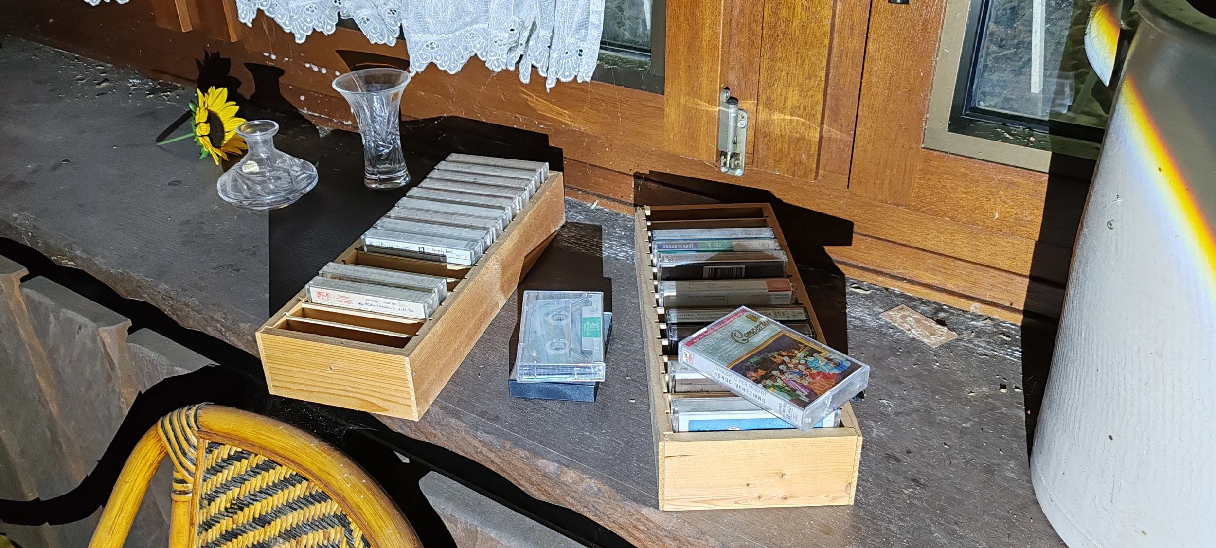 Auf einem Tisch liegen Kassetten, einige in Holzboxen und eine Kassette außerhalb. Es gibt auch zwei Gläser und eine Vase mit Sonnenblume, im Hintergrund eine Holzküchenschrank und eine weiße Wand mit Regenbogenreflexion.