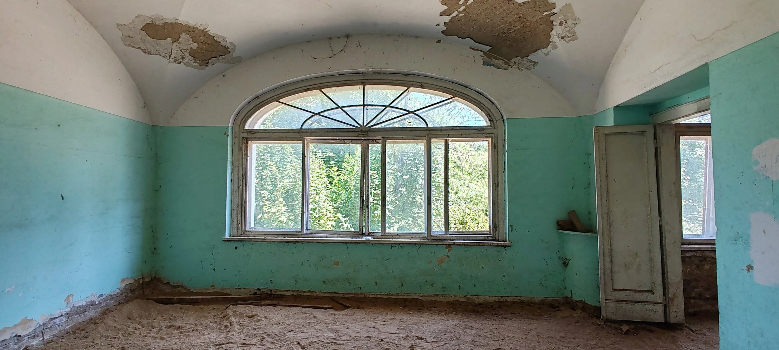 Ein verlassener Raum mit großen fenster, grünen Wänden, Deckenverfall und Sandboden, Blick nach draußen auf Bäume.