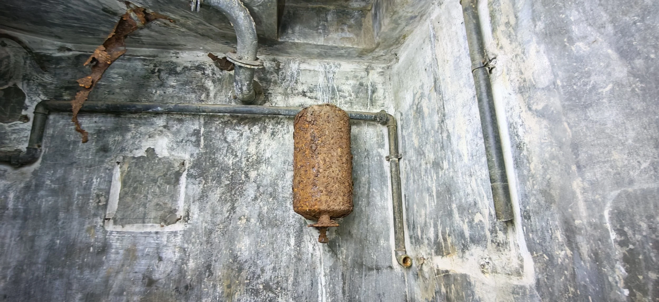 Alte, rostige Wasserleitung an einer verfallenen Zementwand in einem Keller oder Abstellraum.