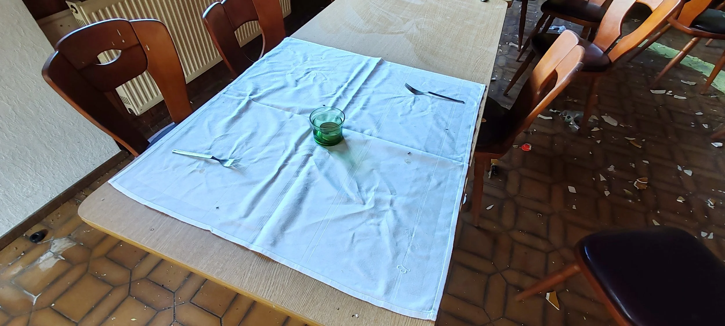 Ein Esstisch mit weißen Tischdecke, auf der eine grüne Glasschale, eine Gabel und ein Messer liegen. Mehrere Holzhocker um den Tisch herum, eine stilvolle, abgenutzte Innenraumumgebung.