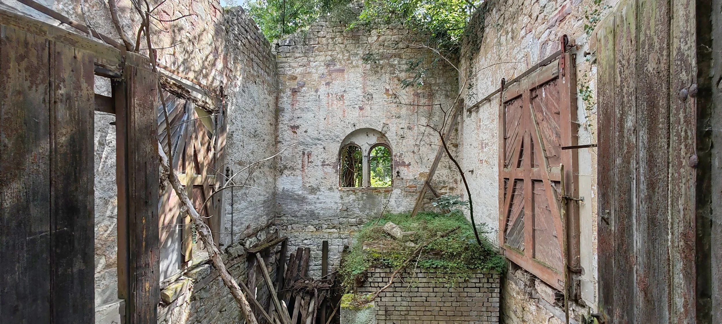 Verfallene, verlassene Ruine mit zerbrochenen Fenstern und verwitterten Holztüren, überwuchert von Pflanzen und Bäumen.