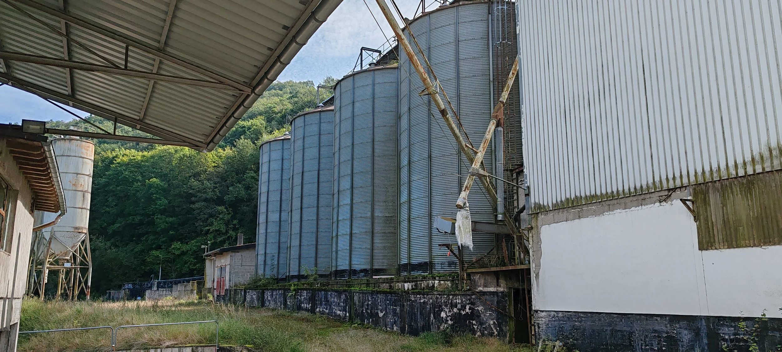 Blick auf alte, verrostete Silos und Fabrikgebäude in einer ländlichen Gegend mit bewaldetem Hügel im Hintergrund.