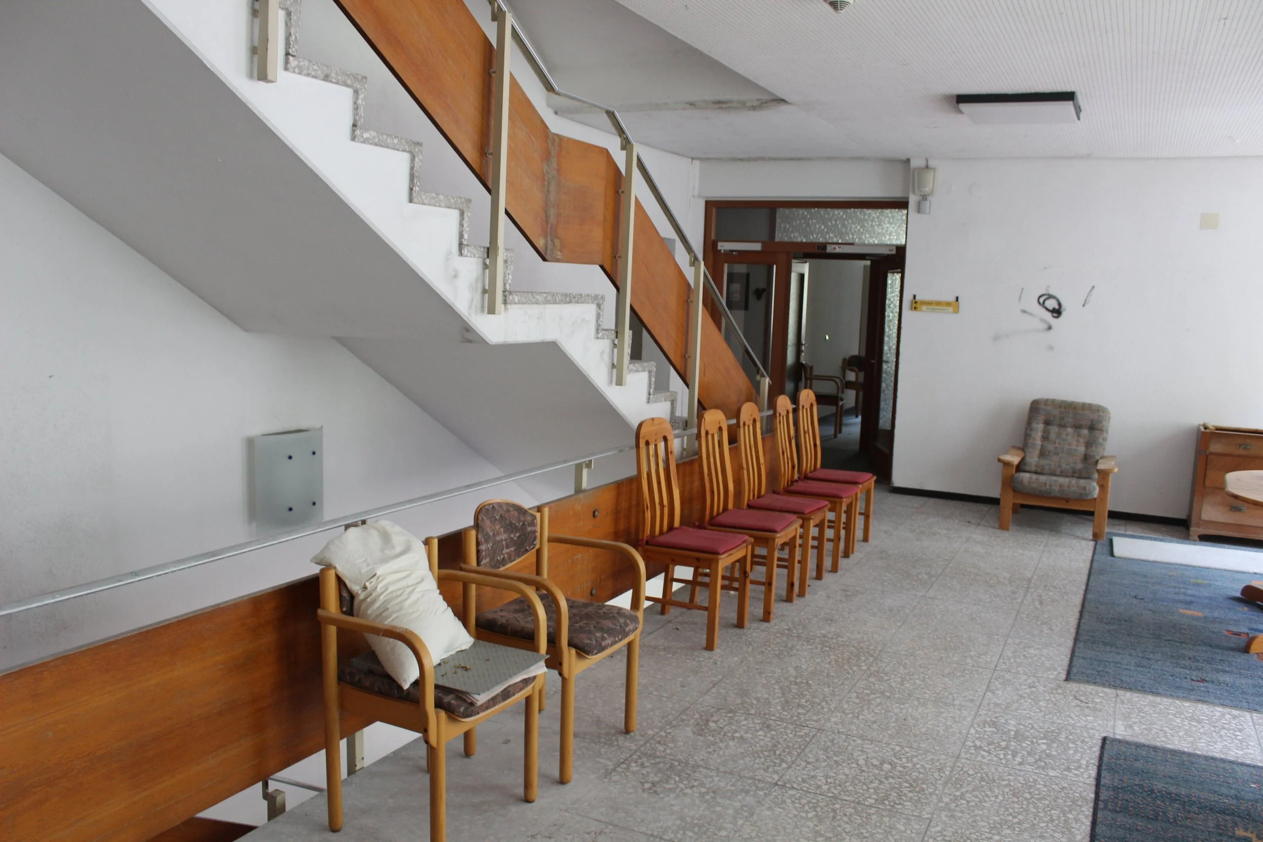 Warteraum mit mehreren Holztischen und Stühlen, einer Treppe im Hintergrund, grauer Teppich, weiße Wände, eine einzelne Bank, eine bequeme Armlehnenstühle und ein gelber Ablagekasten.