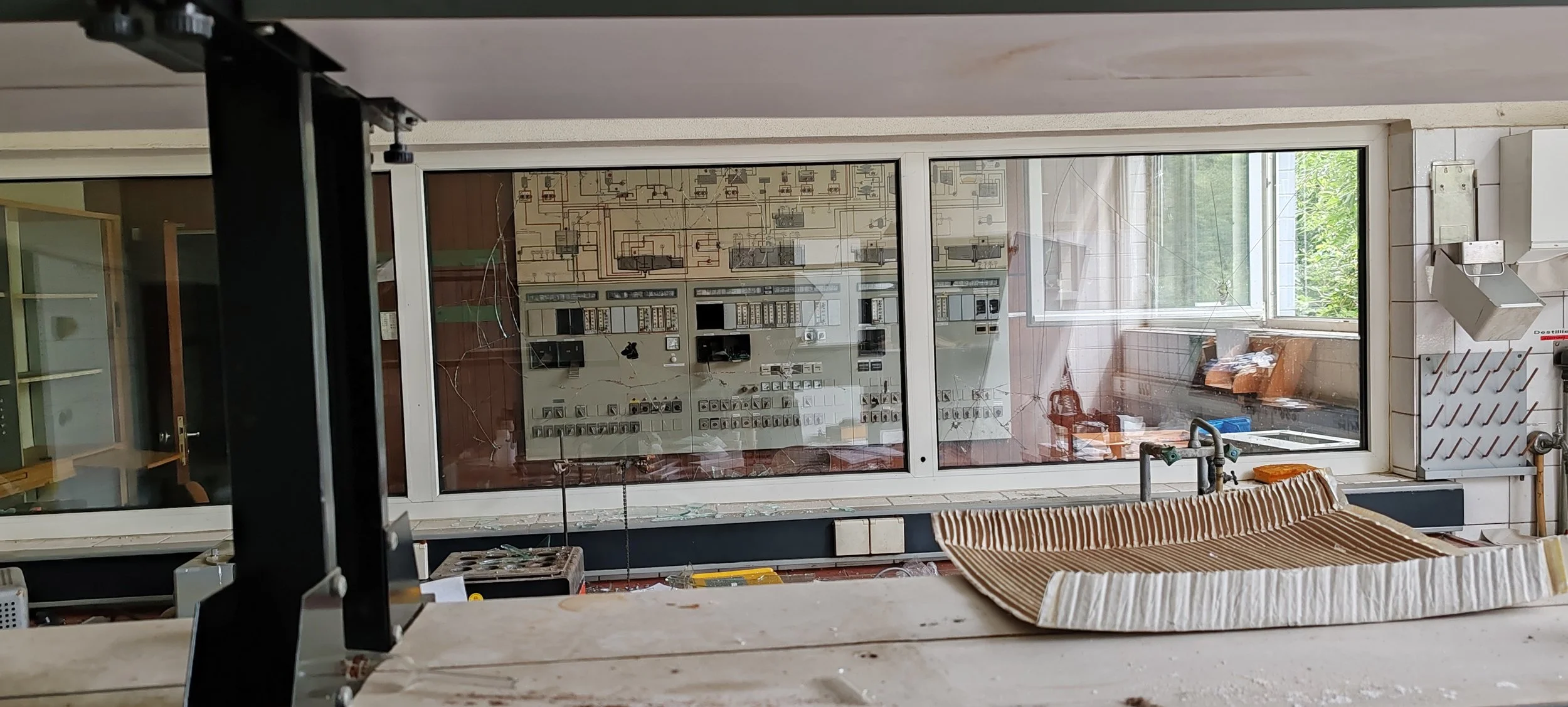 Innenansicht eines alten Elektroniklabors mit einer großen Glasscheibe, die einen Steuerpult mit vielen Schaltern und Schaltkreisen zeigt, und einem Fenster, das auf eine grüne Außenlandschaft blickt.