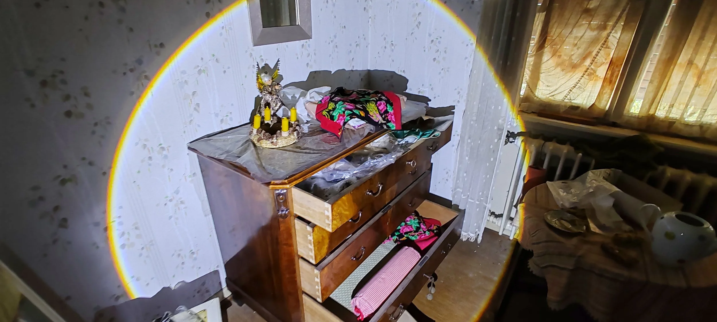 Ein alter Holzkommode mit mehreren Schubladen in einem Zimmer, bedeckt mit Stoffen und Gegenständen, eine Kerzenhalter mit vier Kerzen, ein Altar mit einem Engelstatue, eine Tischdecke mit Mustern, in einem Raum mit älterer Einrichtung, Vorhängen und