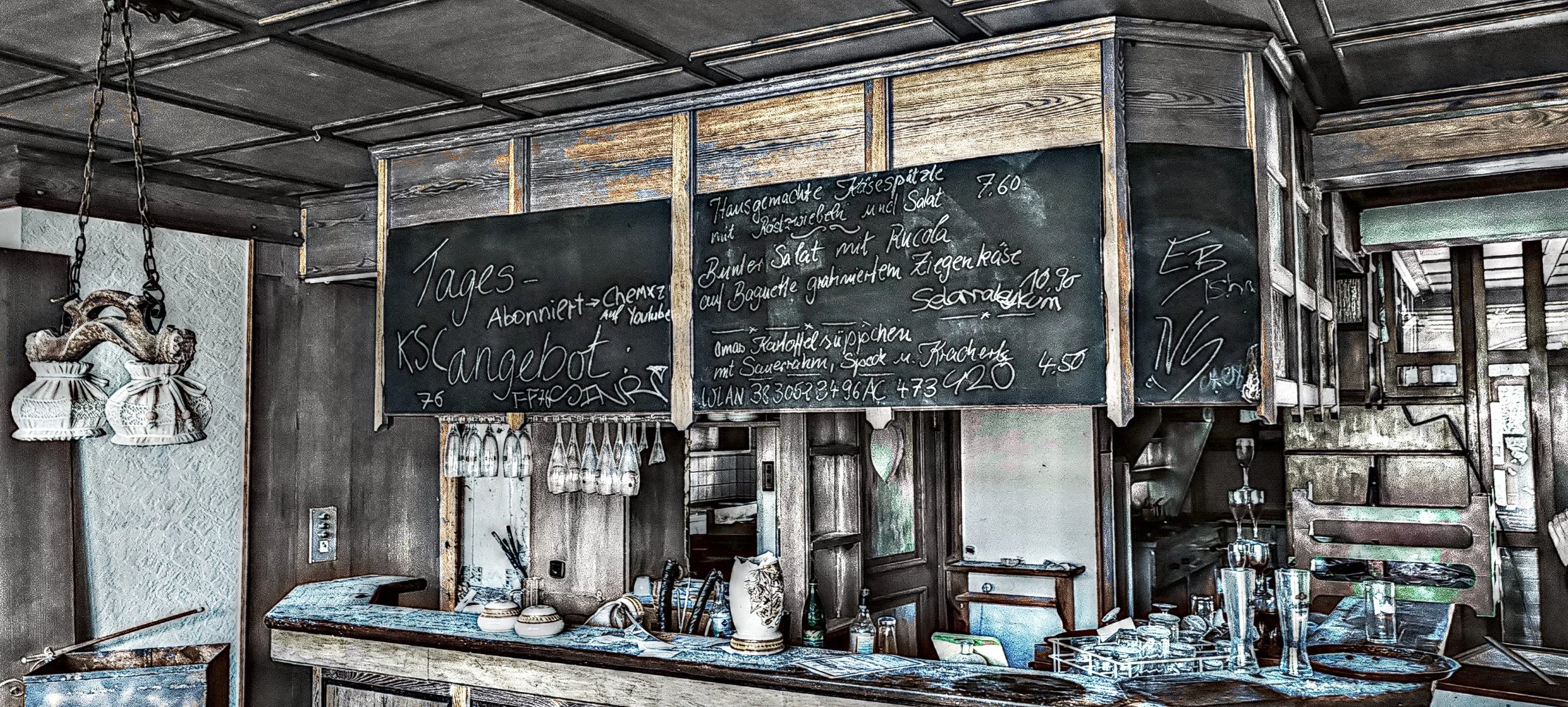 Ein Tresen in einem rustikalen Restaurant mit dunklem Holz und einer schwarzen Tafel mit Speisekarten in deutscher Sprache. Es hängen Weingläser unter der Tafel, und auf dem Tresen stehen Geschirr und Gläser.