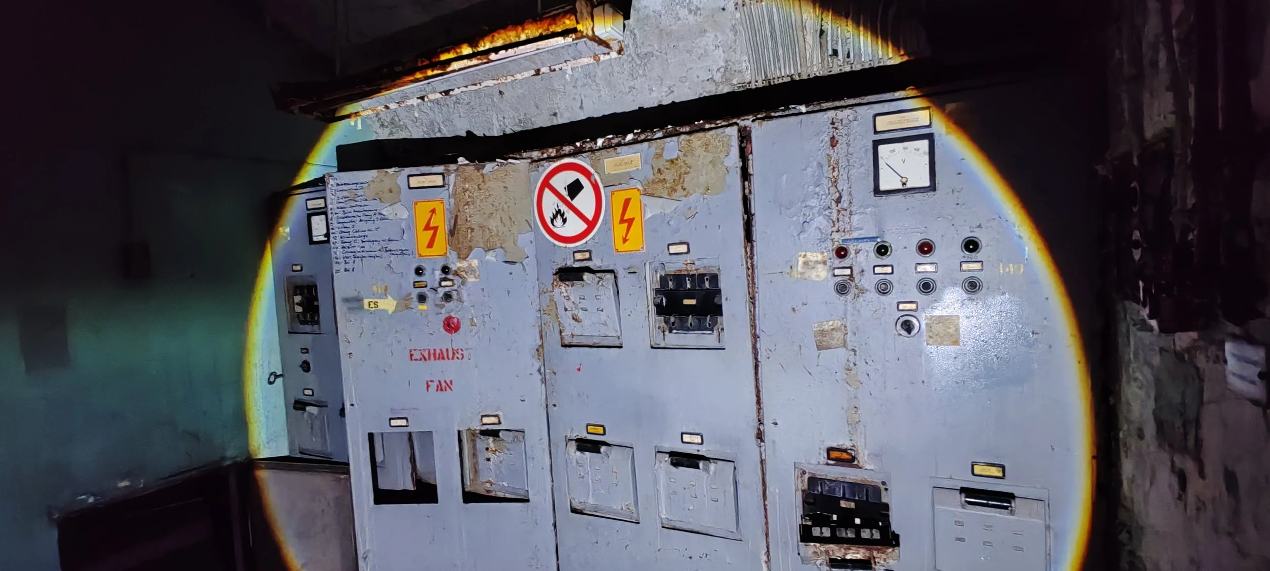 Veralteter, rostiger elektrischer Schaltschrank mit Warnhinweisen und Kontrollanzeigen, in einer dunklen Umgebung, beleuchtet durch eine Taschenlampe.