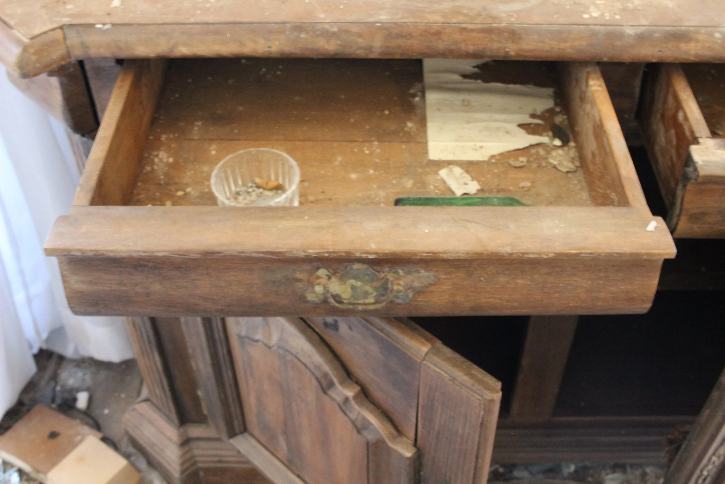 Aufgerissene alte Holzschublade eines Schreibtischs, verschmutzt und beschädigt, mit Staub und kleinen Gegenständen im Inneren.