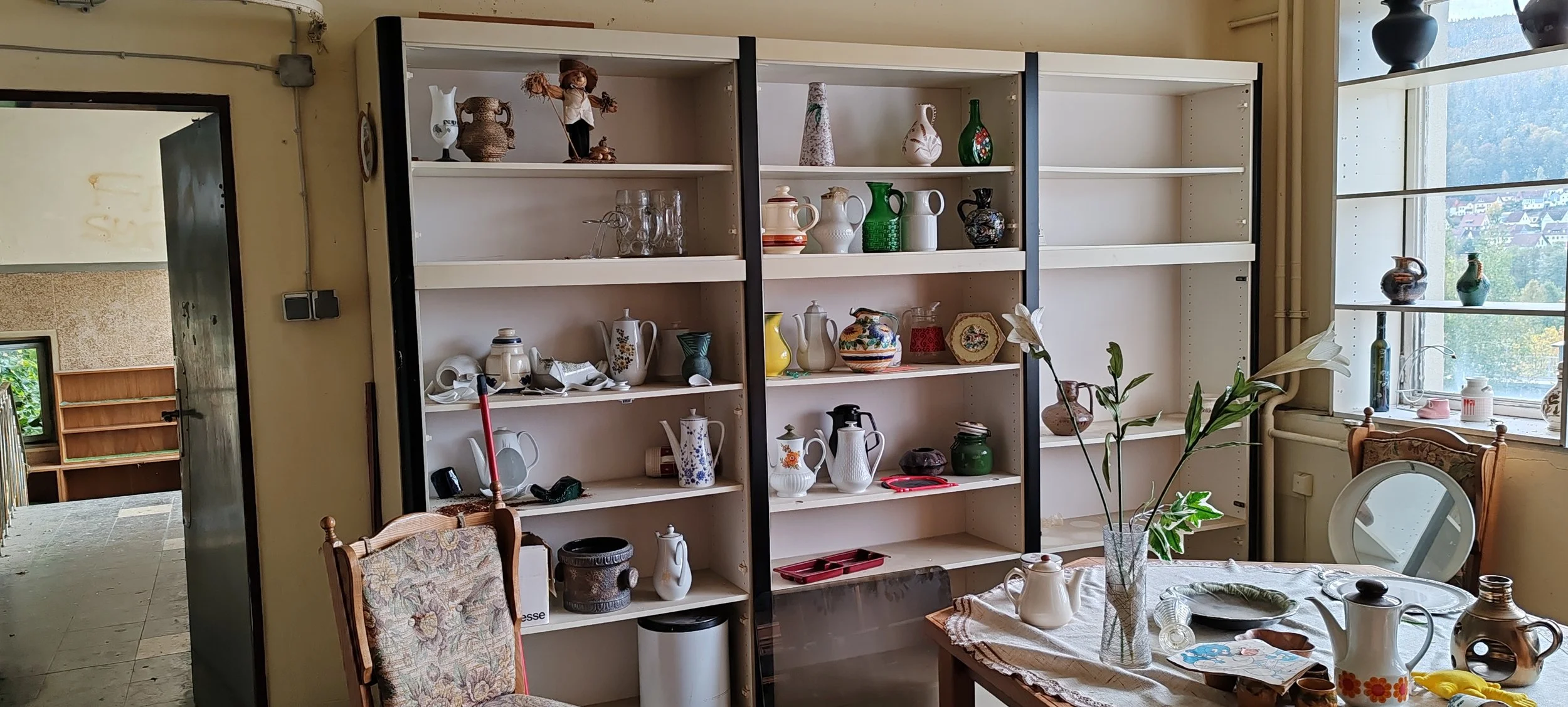 Leeres Bücherregal mit einzelnen Vasen, Geschirr und Dekorationsgegenständen, ein Tisch mit Blumen in einer Vase, auf dem Tisch befinden sich auch Tassen und Porzellan, ein Fenster mit Blick nach draußen.